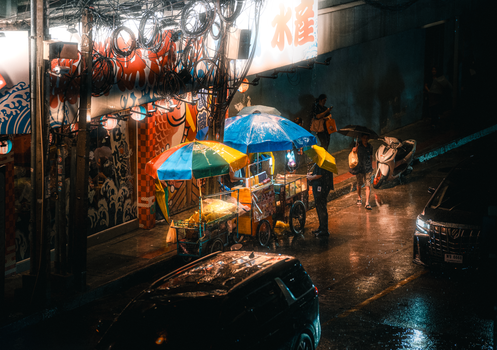 Lively street food stalls under colorful umbrellas in rainy Bangkok at night.
