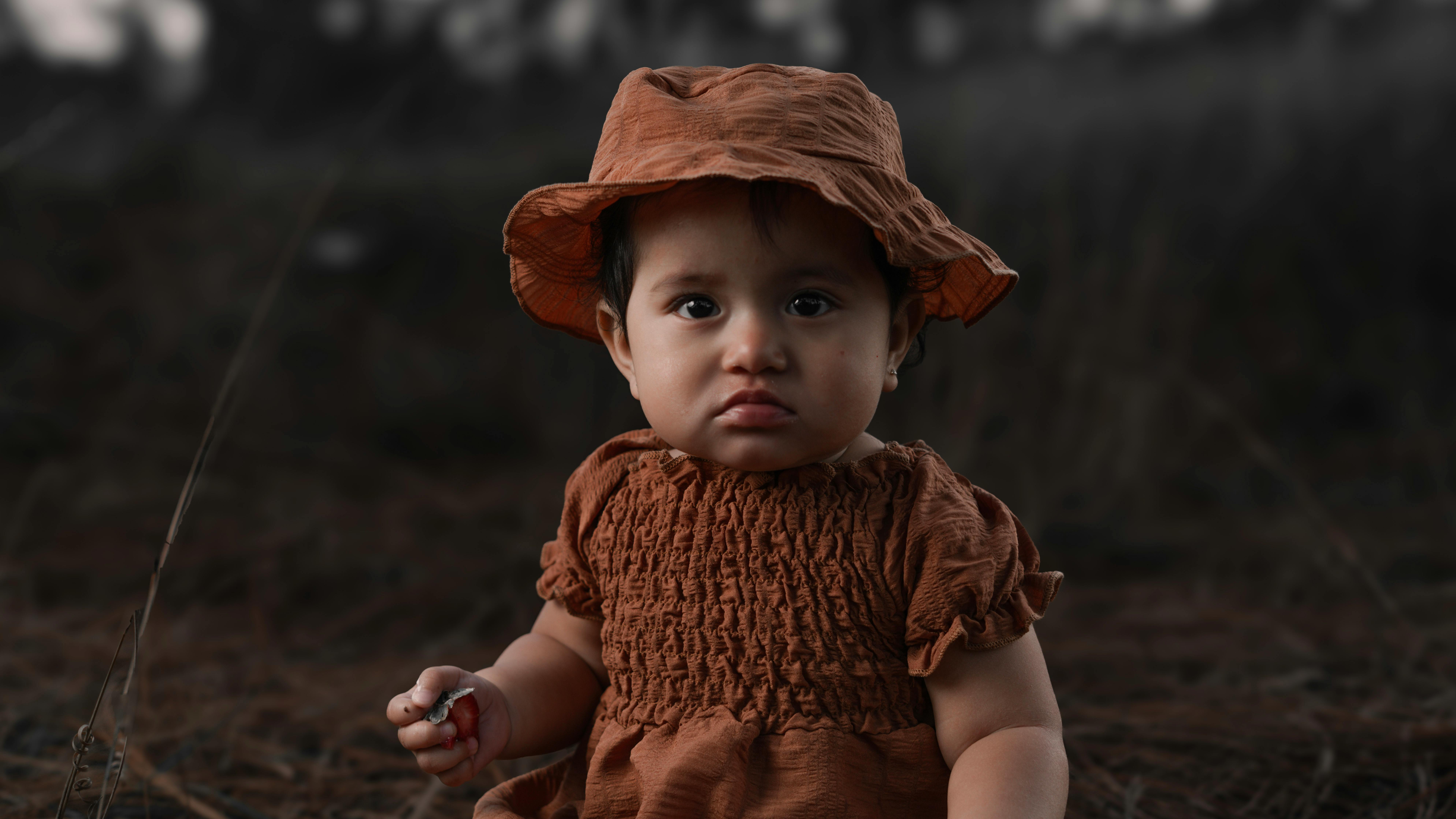 ฟรี เด็กหญิงตัวน้อยน่ารักสวมชุดและหมวกสีน้ำตาล นั่งอยู่ท่ามกลางธรรมชาติ สร้างภาพถ่ายกลางแจ้งที่สงบสุข คลังภาพถ่าย