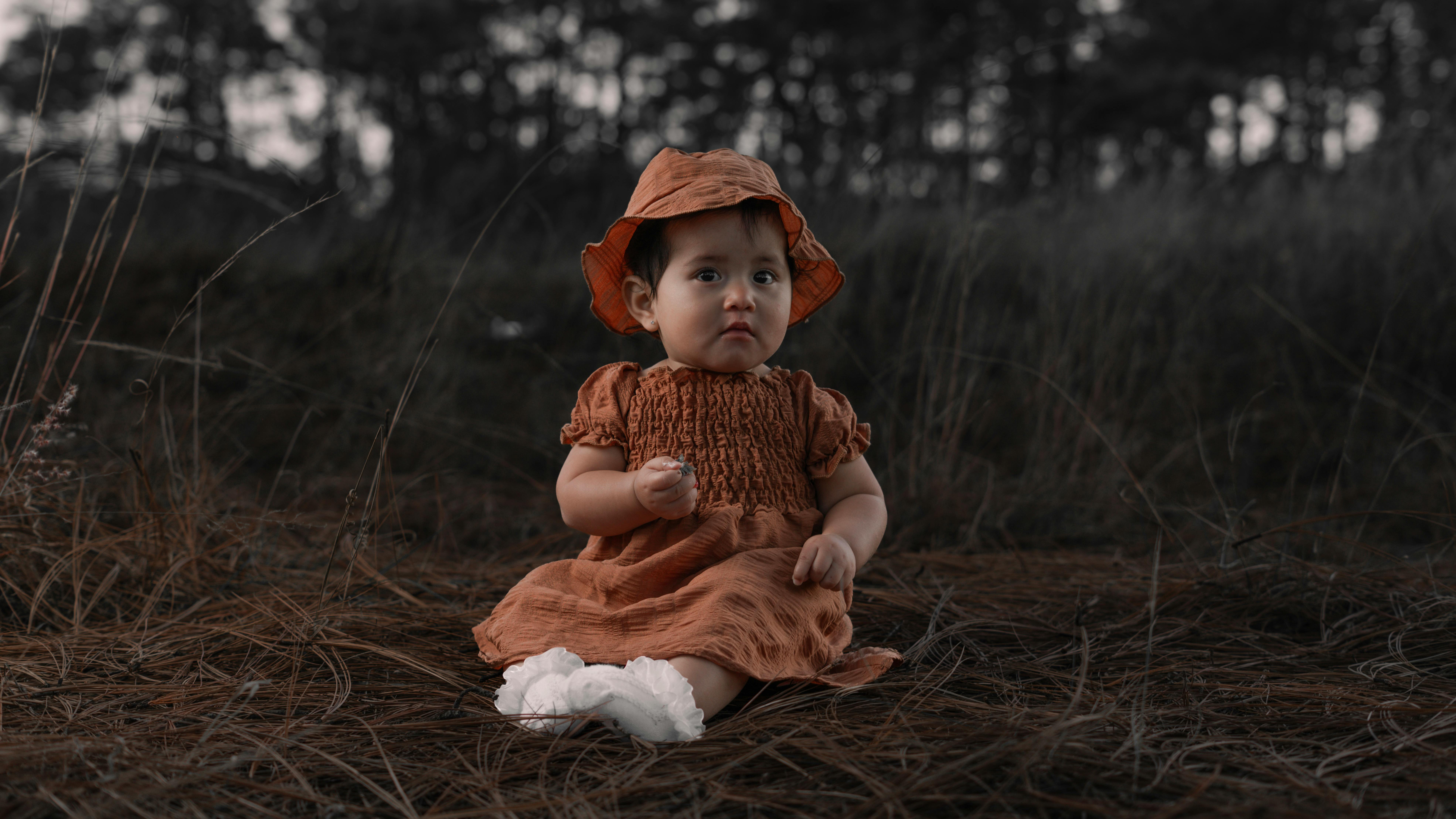 Kostenlos Baby sitzt mit orangefarbenem Kleid und Hut auf braunem Gras in herbstlicher Umgebung. Stock-Foto