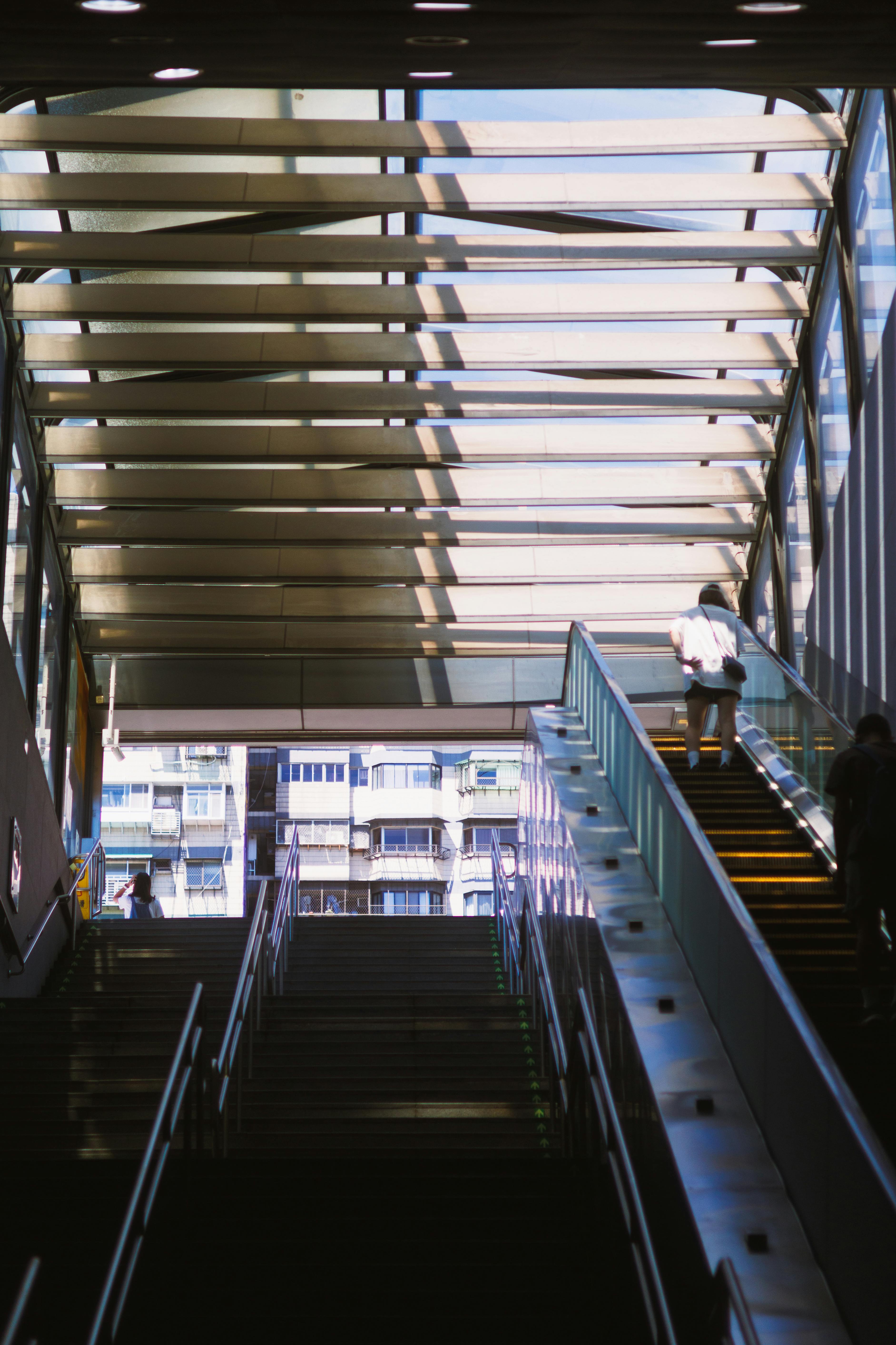 Gratis Una persona subiendo una escalera mecánica en el interior de una estación urbana iluminada por el sol, con sombras y diseños geométricos. Foto de stock