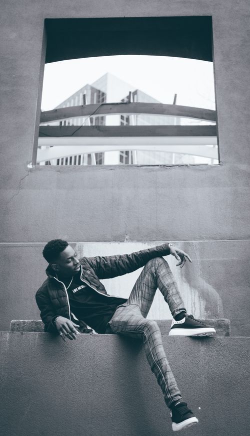 Free Black and white portrait of a fashionable man posing on a city ledge with modern architecture in the background. Stock Photo