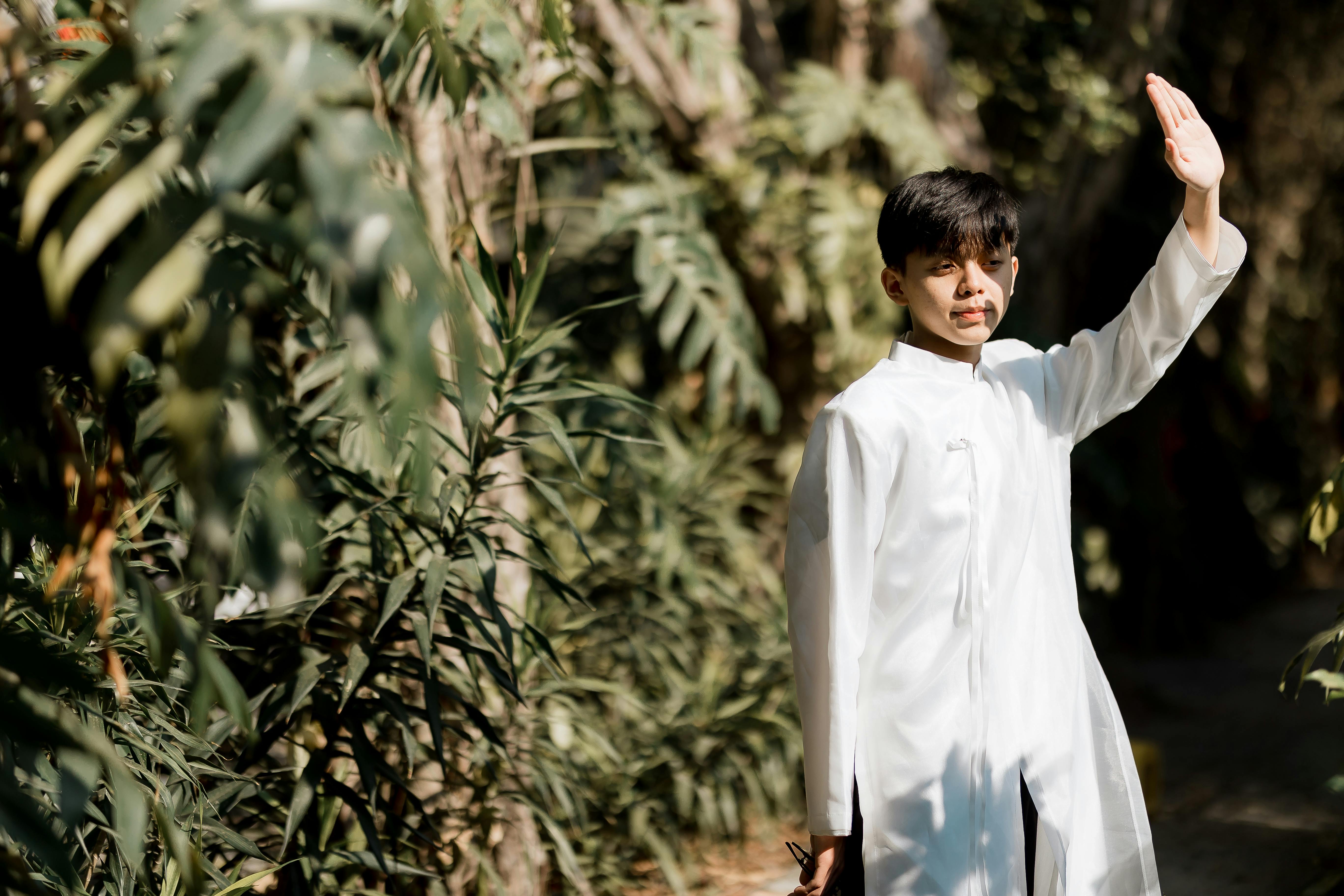 Gratis Seorang pemuda mengenakan pakaian tradisional putih melambaikan tangan sambil berdiri di luar ruangan yang dikelilingi oleh tanaman hijau yang rimbun. Foto Stok