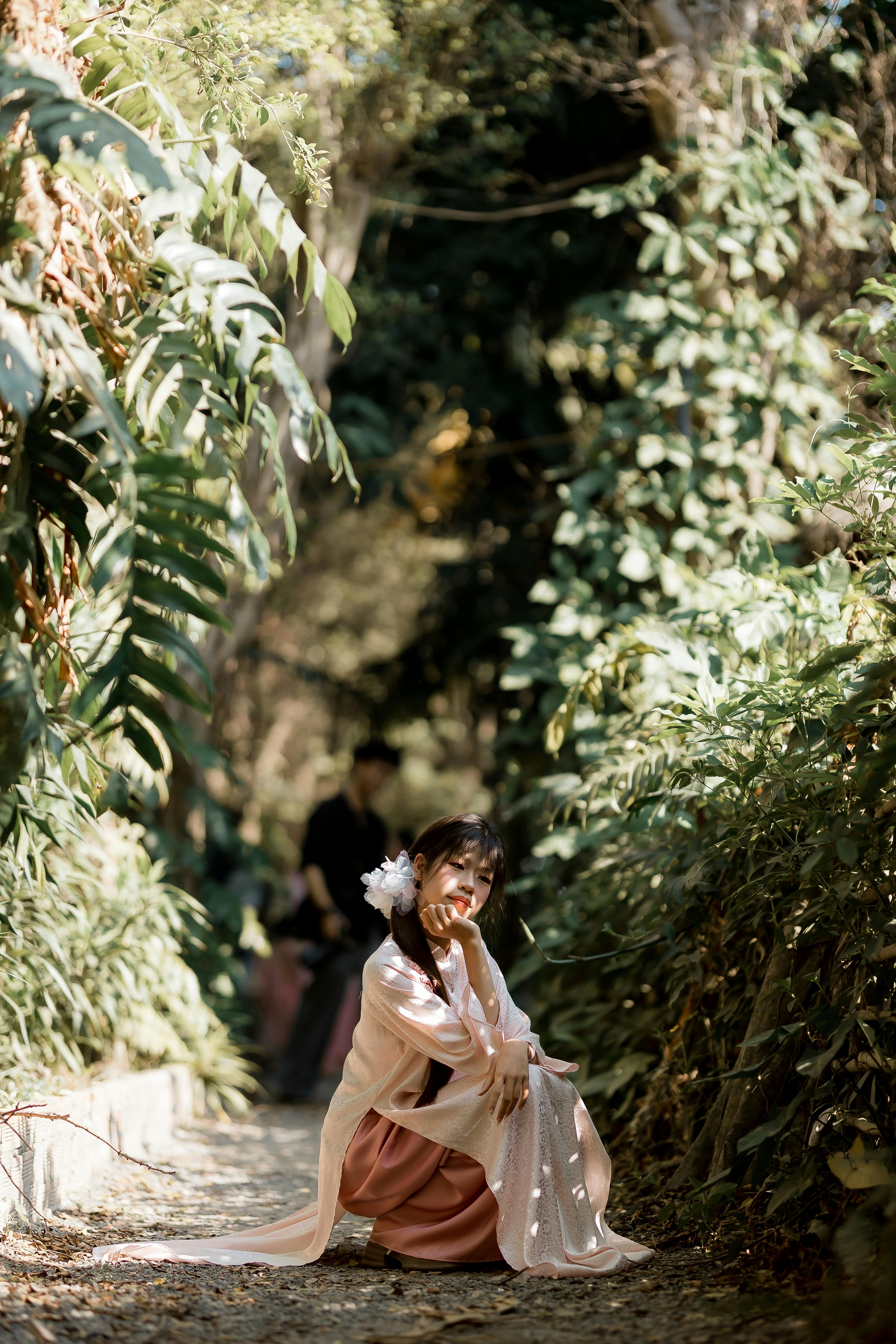 Gratis Seorang wanita muda yang elegan berpose anggun di jalan setapak taman yang diterangi sinar matahari, dikelilingi oleh tanaman hijau yang rimbun. Foto Stok