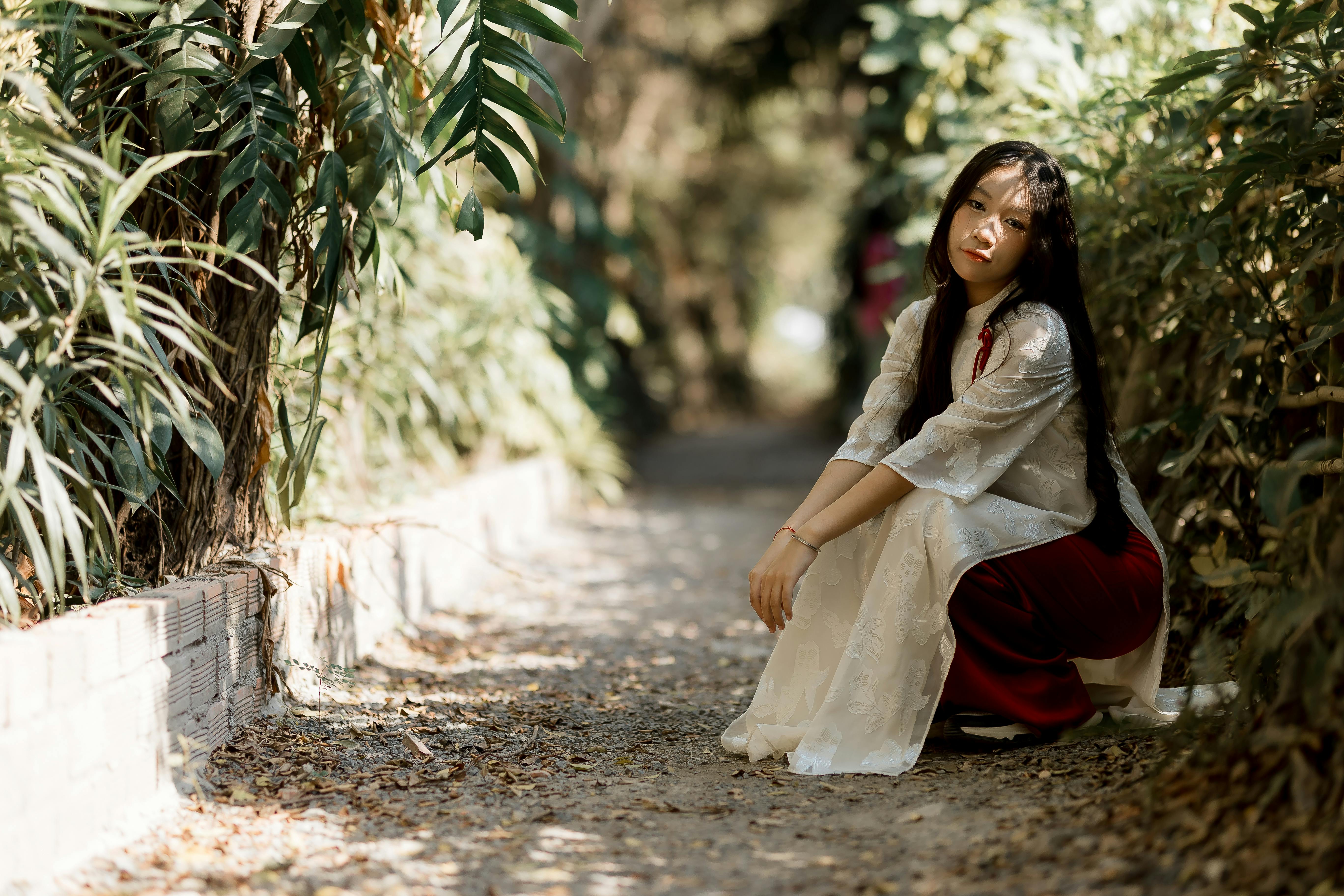 ingyenes Egy hagyományos öltözéket viselő nő ül egy napsütötte ösvényen, buja növényzettel körülvéve, nyugalmat sugározva. Stockfotó