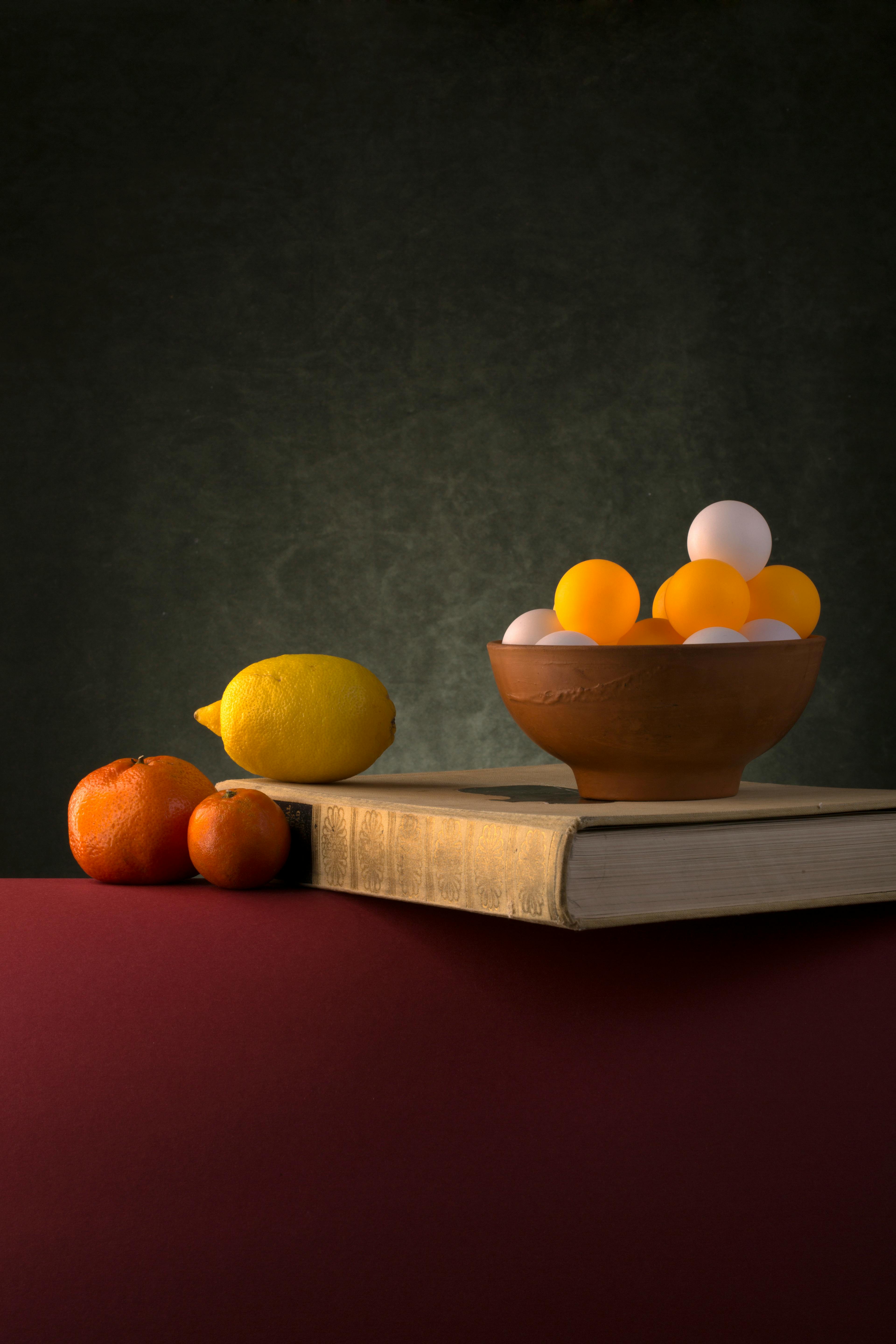 Free A conceptual still life featuring a bowl of spheres, fruit, and a book on a dark textured background. Stock Photo