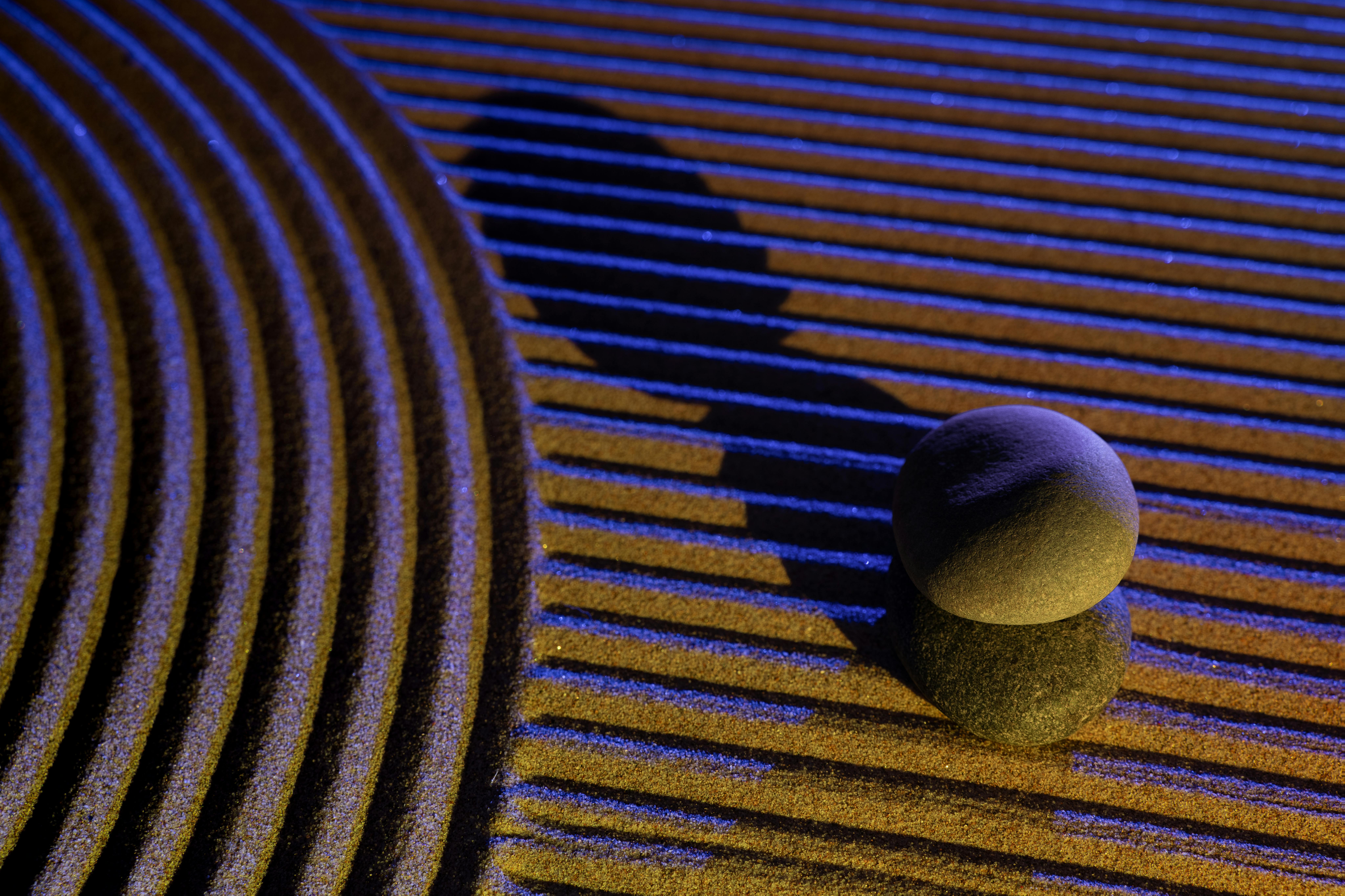 Kostnadsfria Lugn och balanserad komposition av zenstenar med blått ljus som kastar skuggor på texturerad sand. Stock foto