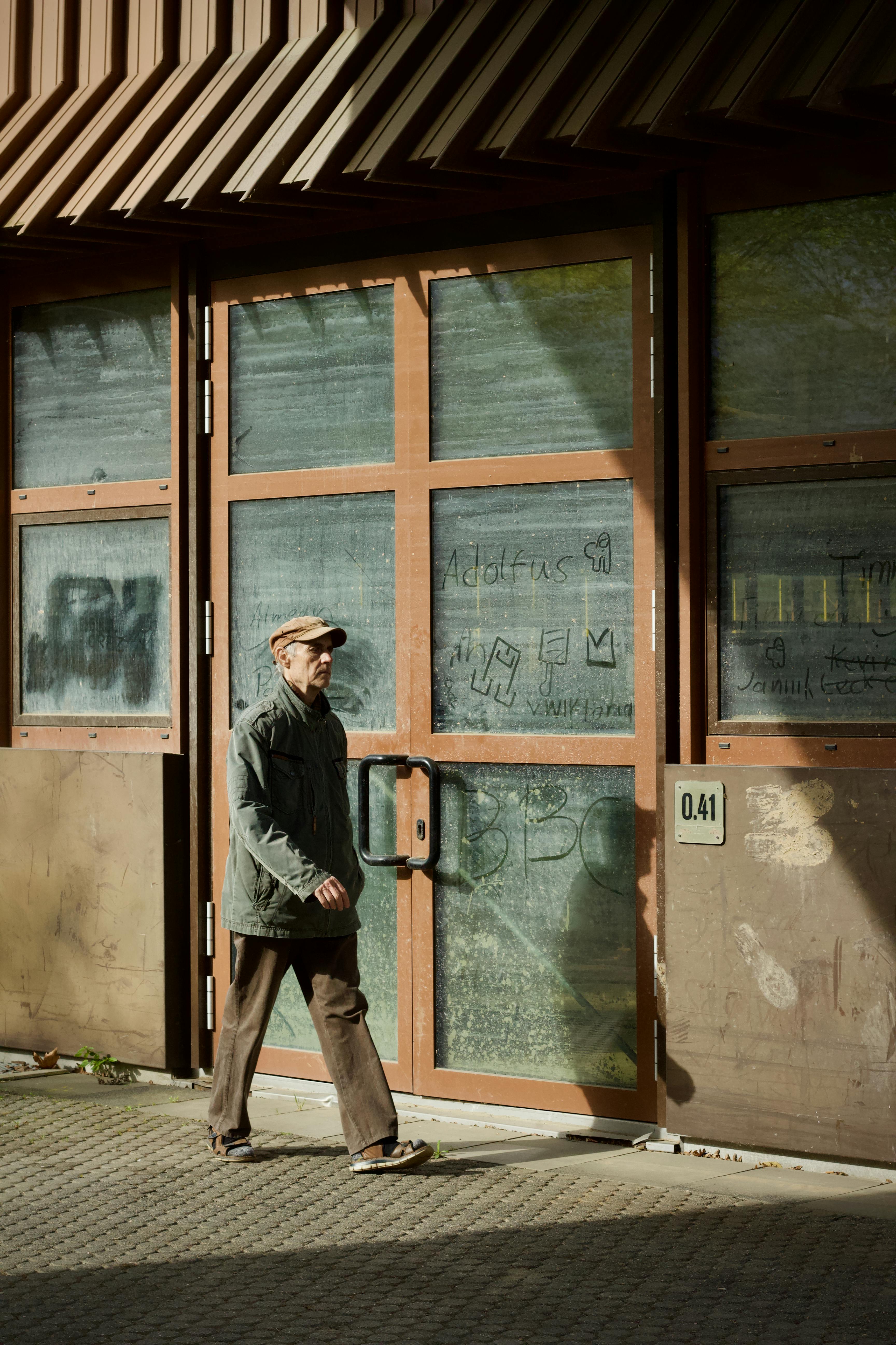 Δωρεάν Ηλικιωμένος άνδρας περπατάει δίπλα από ένα μεγάλο βιομηχανικό κτίριο με γκράφιτι και ηλιακό φως να ρίχνουν σκιές. Φωτογραφία από στοκ φωτογραφιών