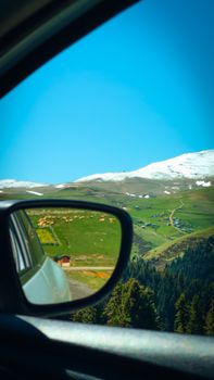 View of snow-capped mountains and green valleys from a car mirror on a sunny day.