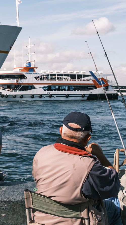 Free Elderly man fishing by a harbor, views of a passenger ferry on a sunny day. Stock Photo
