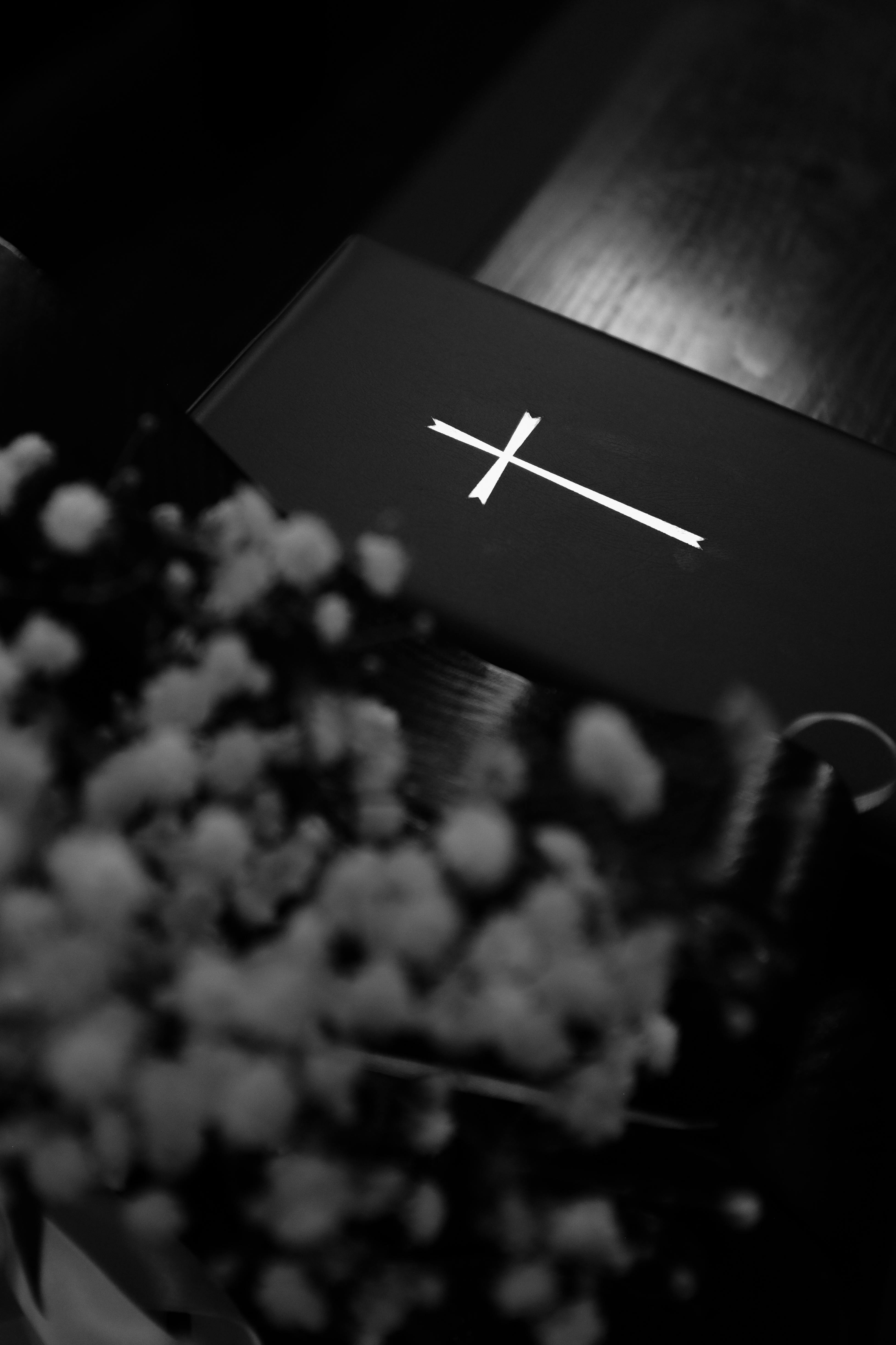 Free A monochrome image featuring a cross on a Bible with blurred flowers in the foreground. Stock Photo