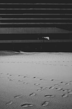 Monochrome image of footprints on a sandy beach in front of a modern building under cloudy skies.