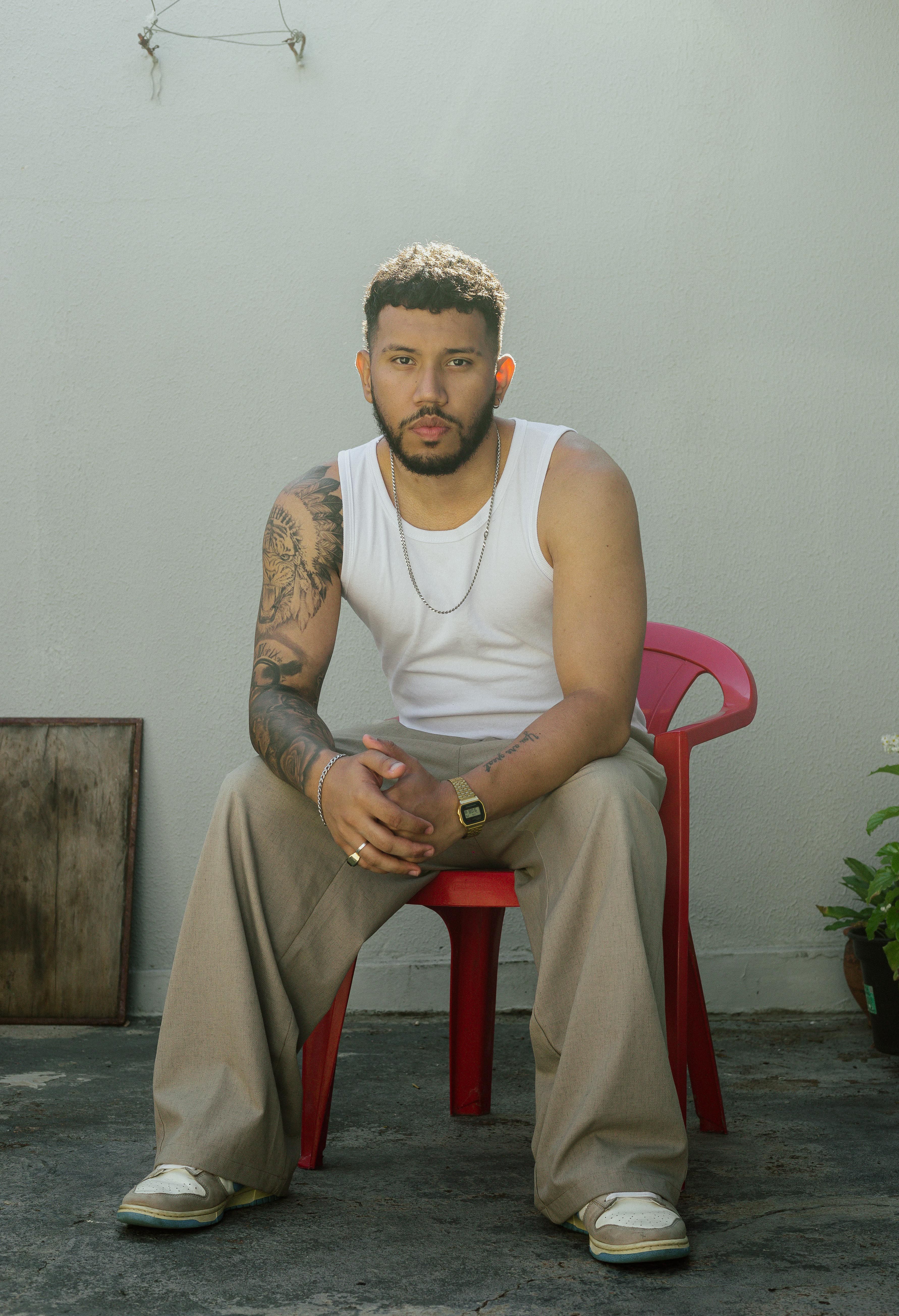 Relaxed man with tattoos seated in a yard, natural lighting, São Paulo.