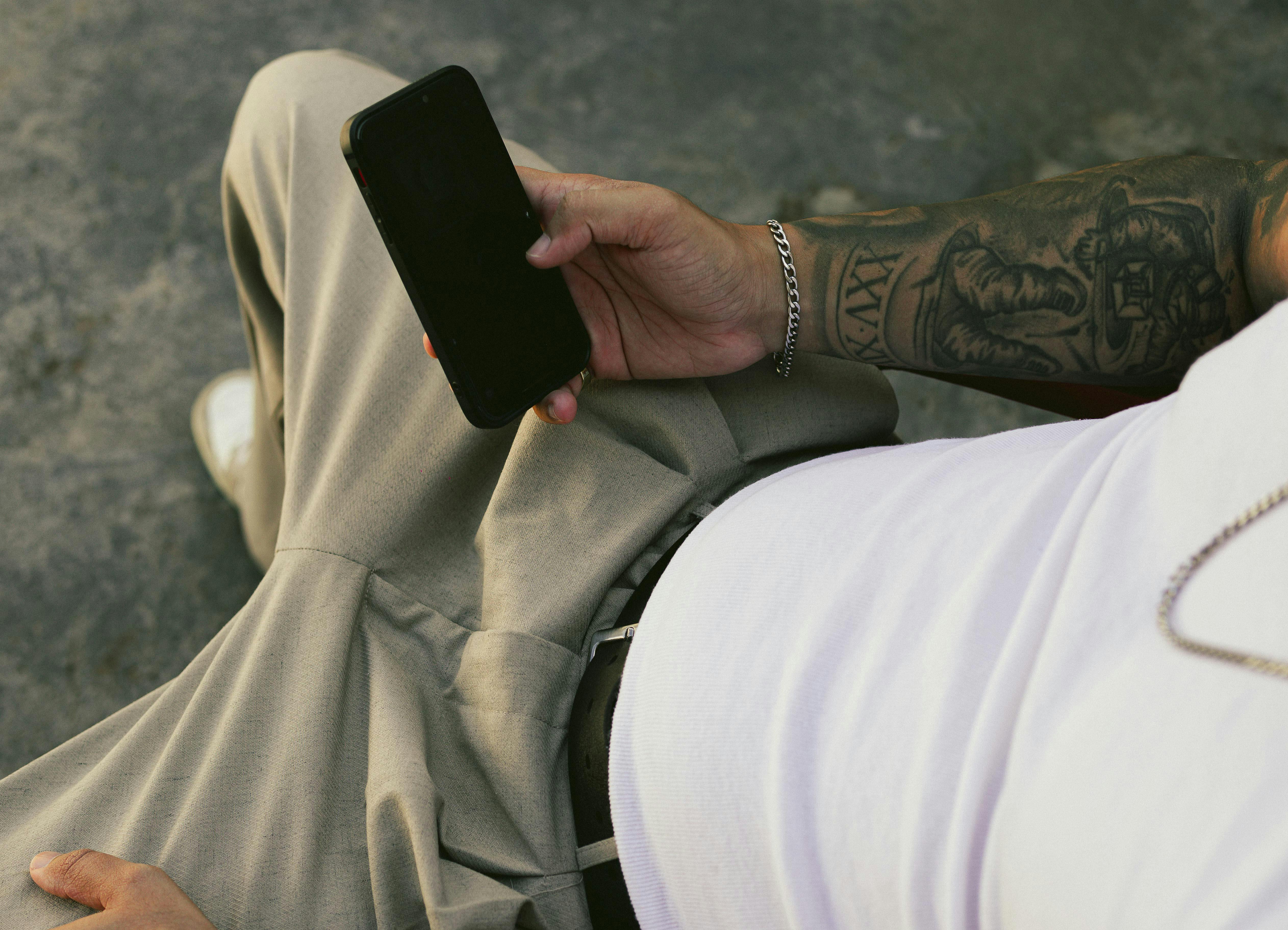 Free Close-up of a tattooed arm holding a smartphone, showcasing urban casual style. Stock Photo