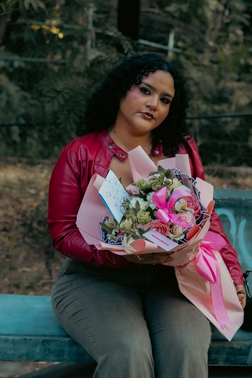 Free Portrait of a woman in a red jacket holding a flower bouquet, sitting on an outdoor bench. Stock Photo