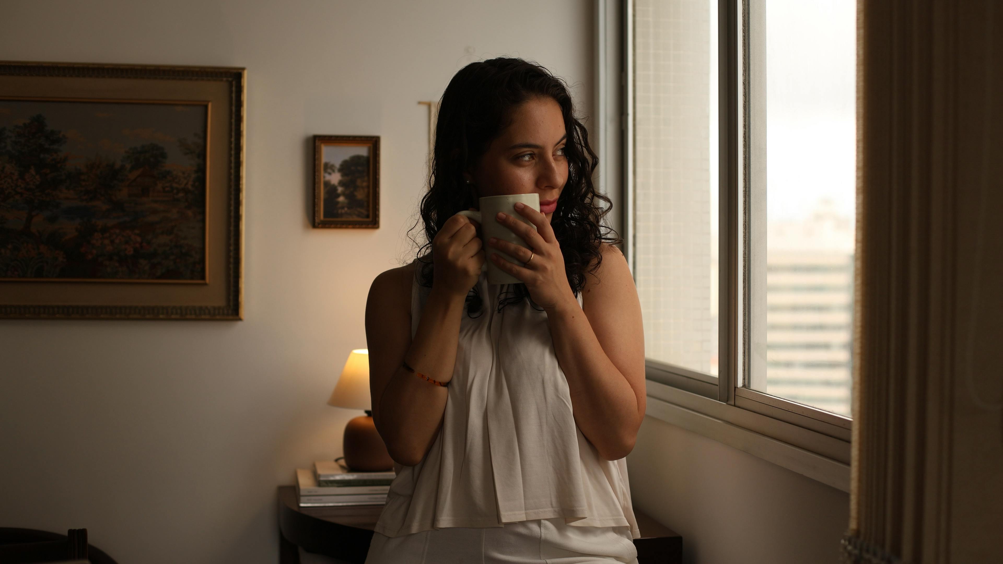 Young woman gazing out window with coffee, enjoying morning indoors.