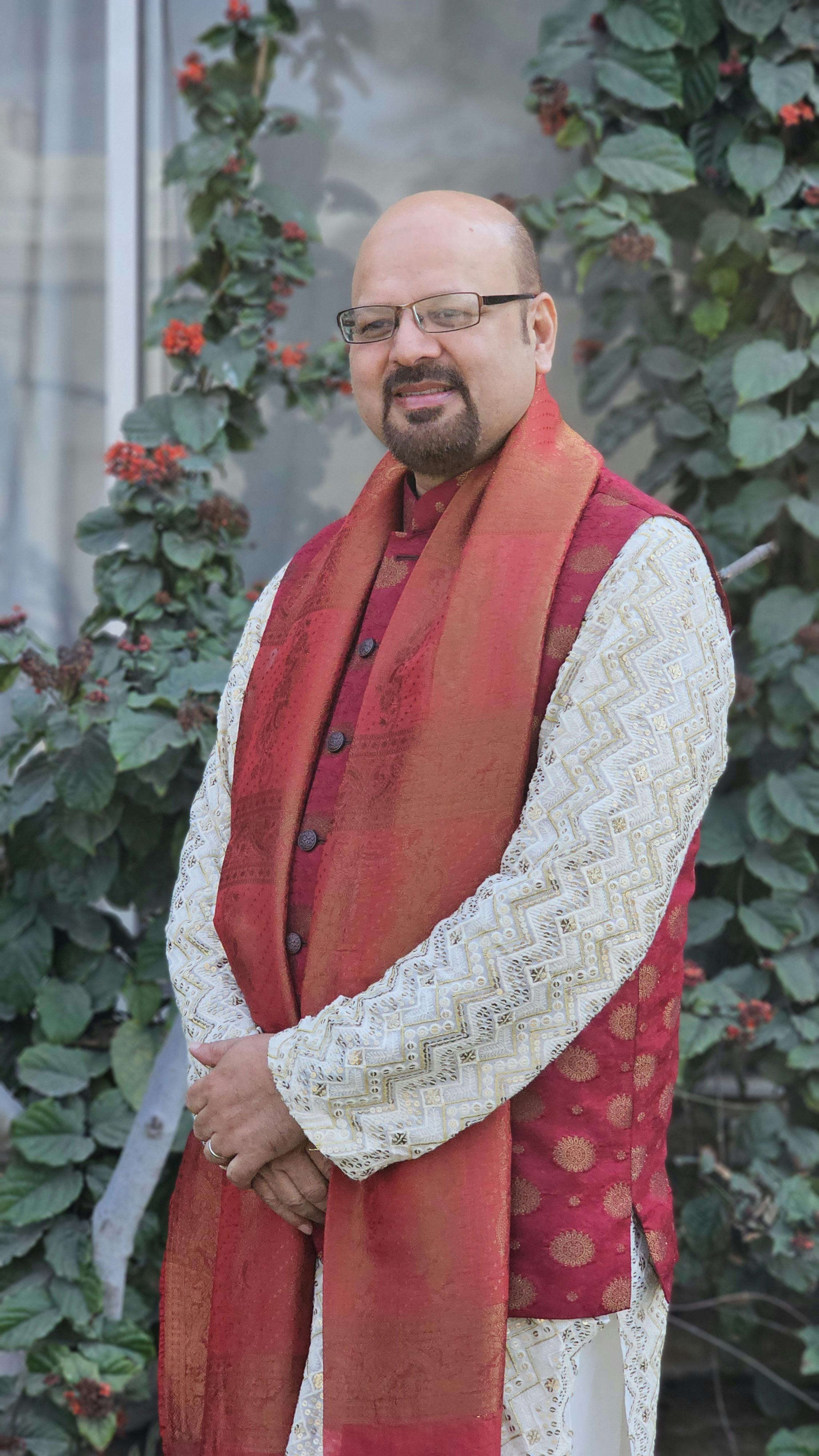 A man in elegant traditional attire poses confidently outdoors with greenery background.