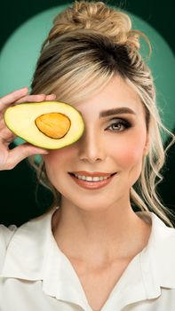 Elegant woman with avocado half over eye on green background, showcasing beauty and wellness.