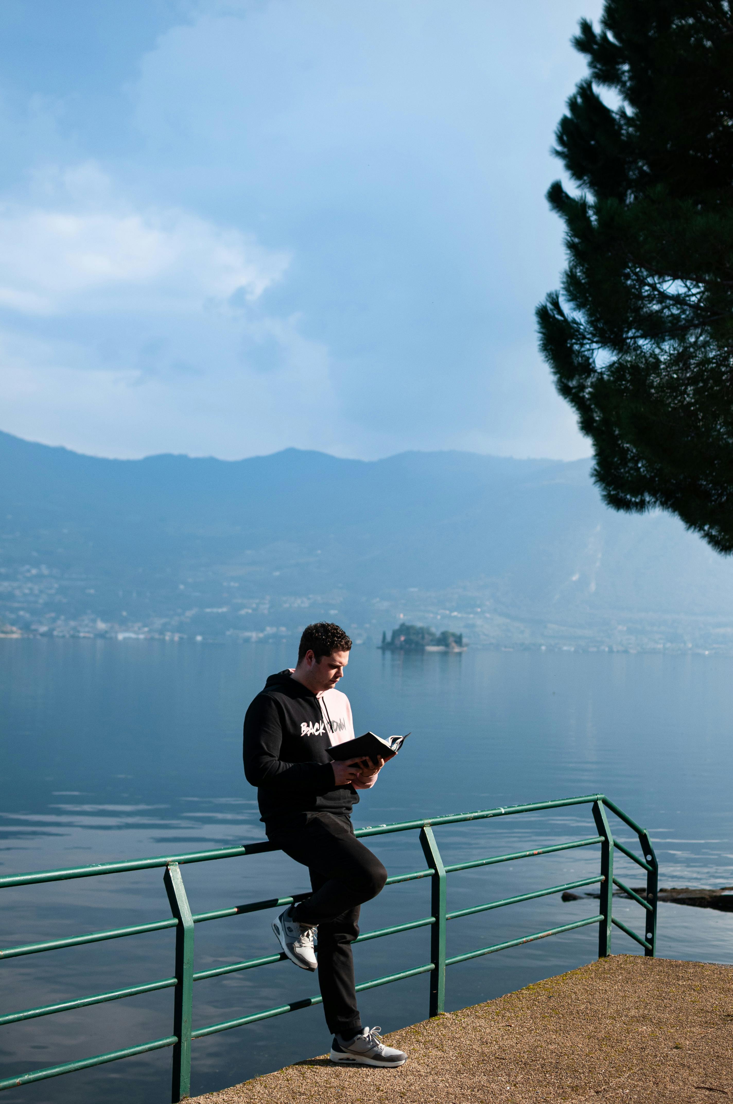 Gratis Un uomo legge un libro sulle rive del placido lago d'Iseo, con le montagne sullo sfondo. Foto a disposizione