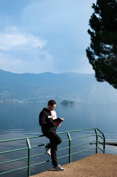 A man reading a book by the serene Lake Iseo with mountains in the background.