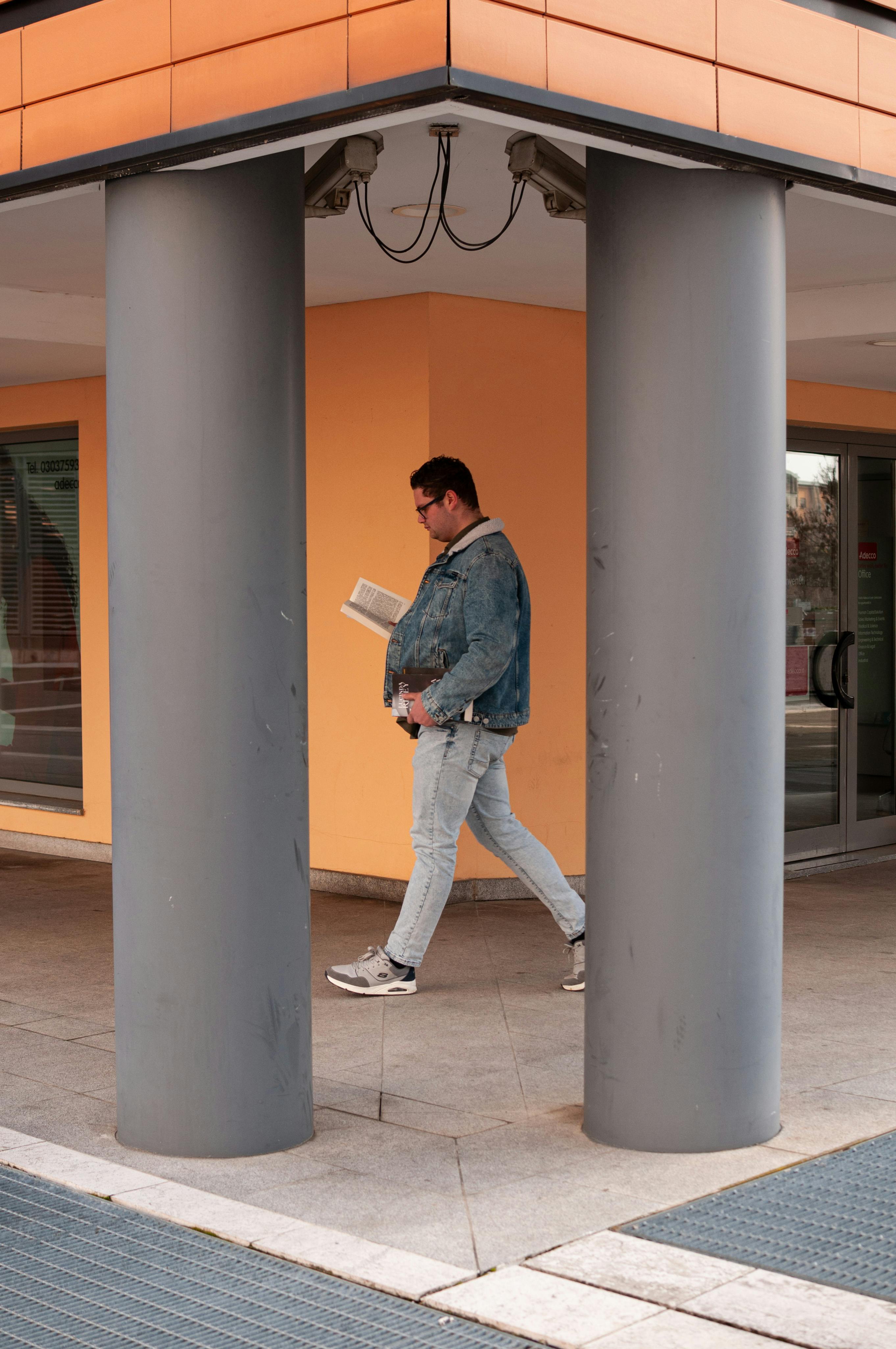 Kostenlos Ein lässig gekleideter Mann liest ein Buch, während er an Säulen in Mailand vorbeigeht. Stock-Foto
