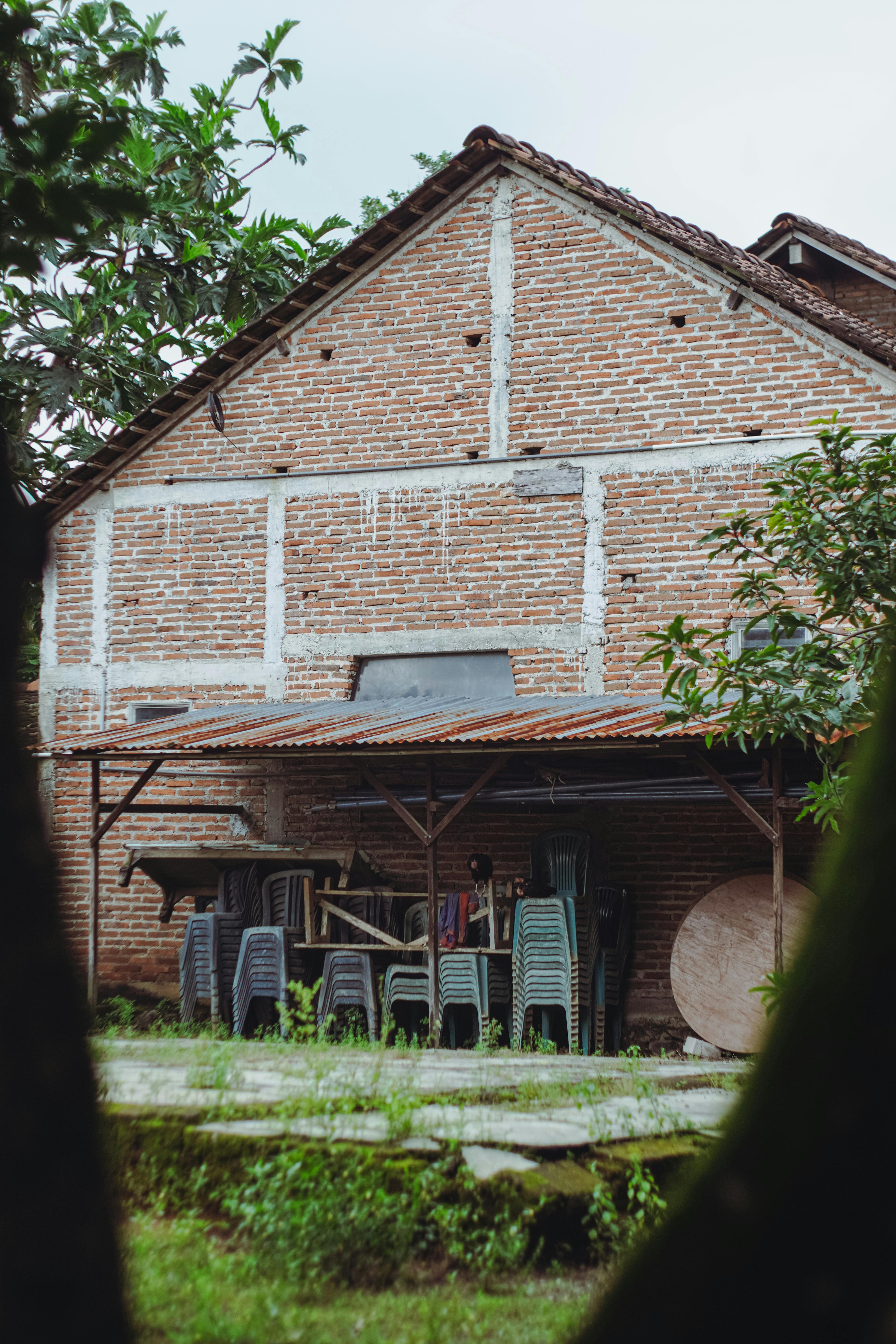 Gratis Una rustica casa in mattoni con tettoia e sedie di plastica impilate in un lussureggiante ambiente esterno. Foto a disposizione