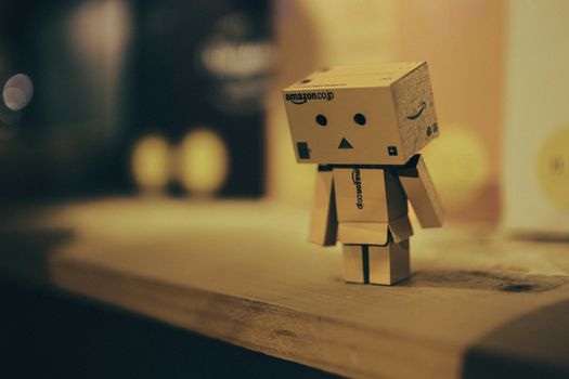 A small cardboard robot figurine sitting on a wooden shelf in a warmly lit room.
