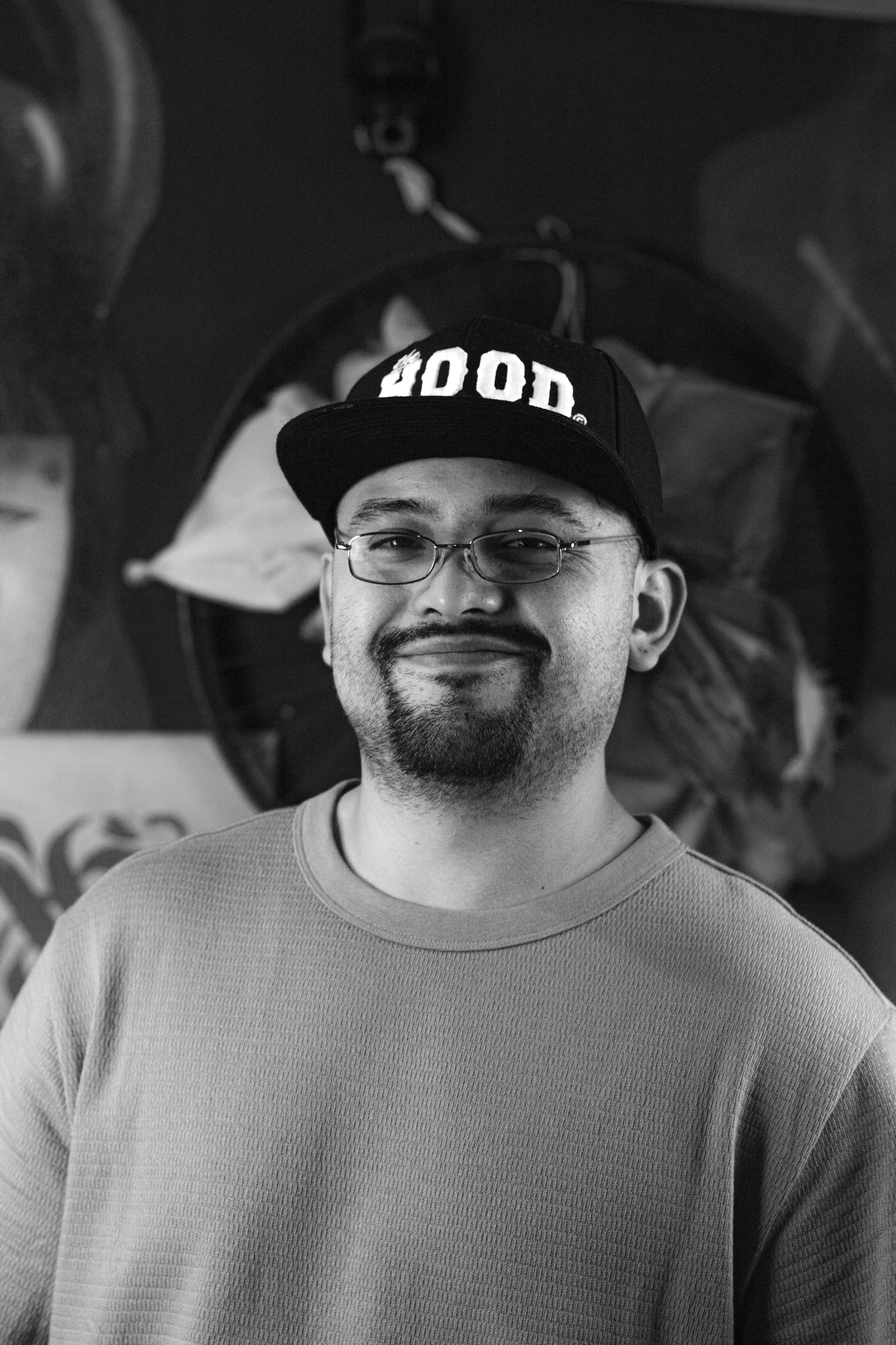 Gratis Uomo sorridente che indossa un berretto con la scritta "HOOD", in piedi in un interno con un'opera d'arte sullo sfondo. Foto a disposizione