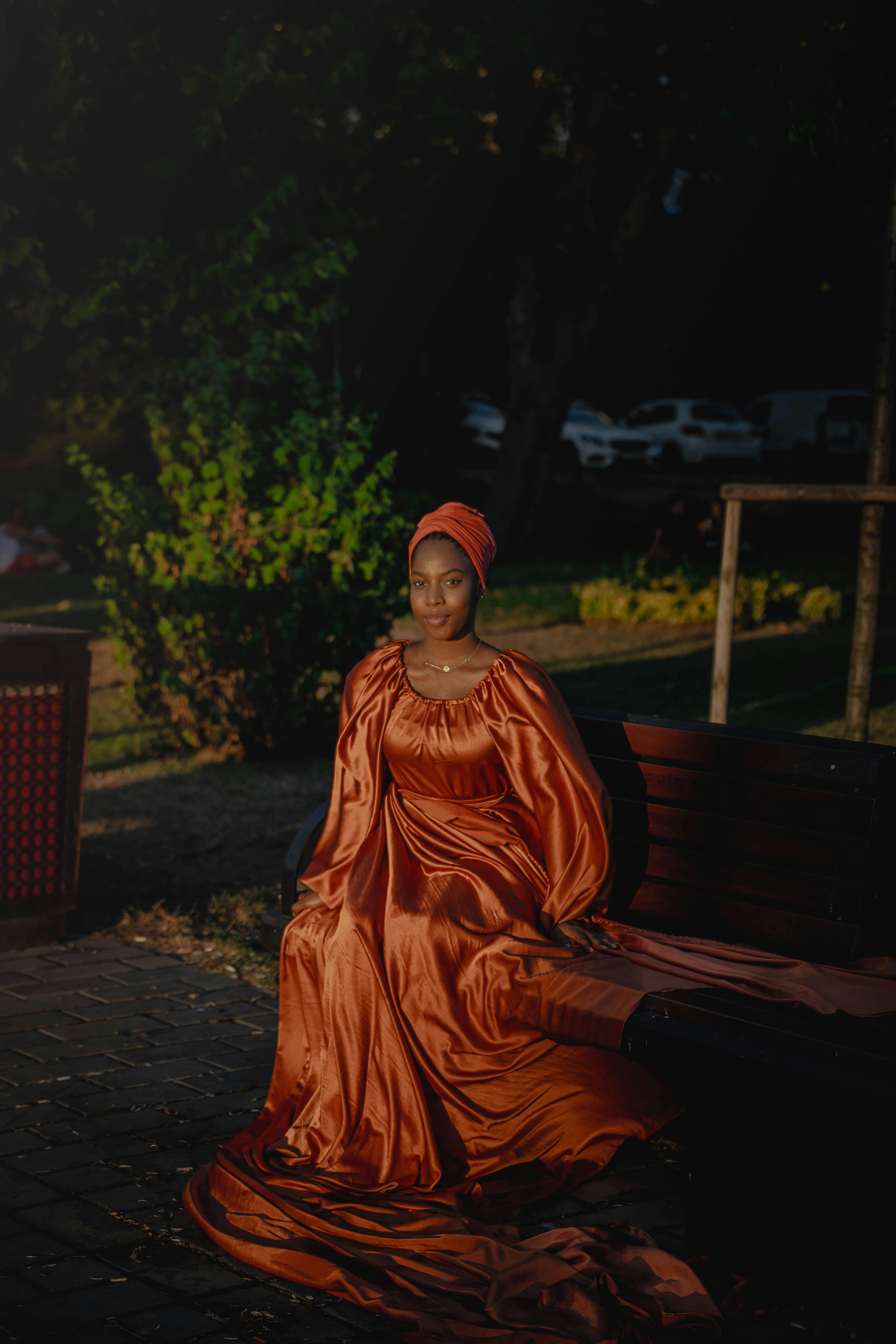 Ilmainen Nainen hulmuavassa oranssissa mekossa istuu penkillä puistossa. Kuvapankkikuva