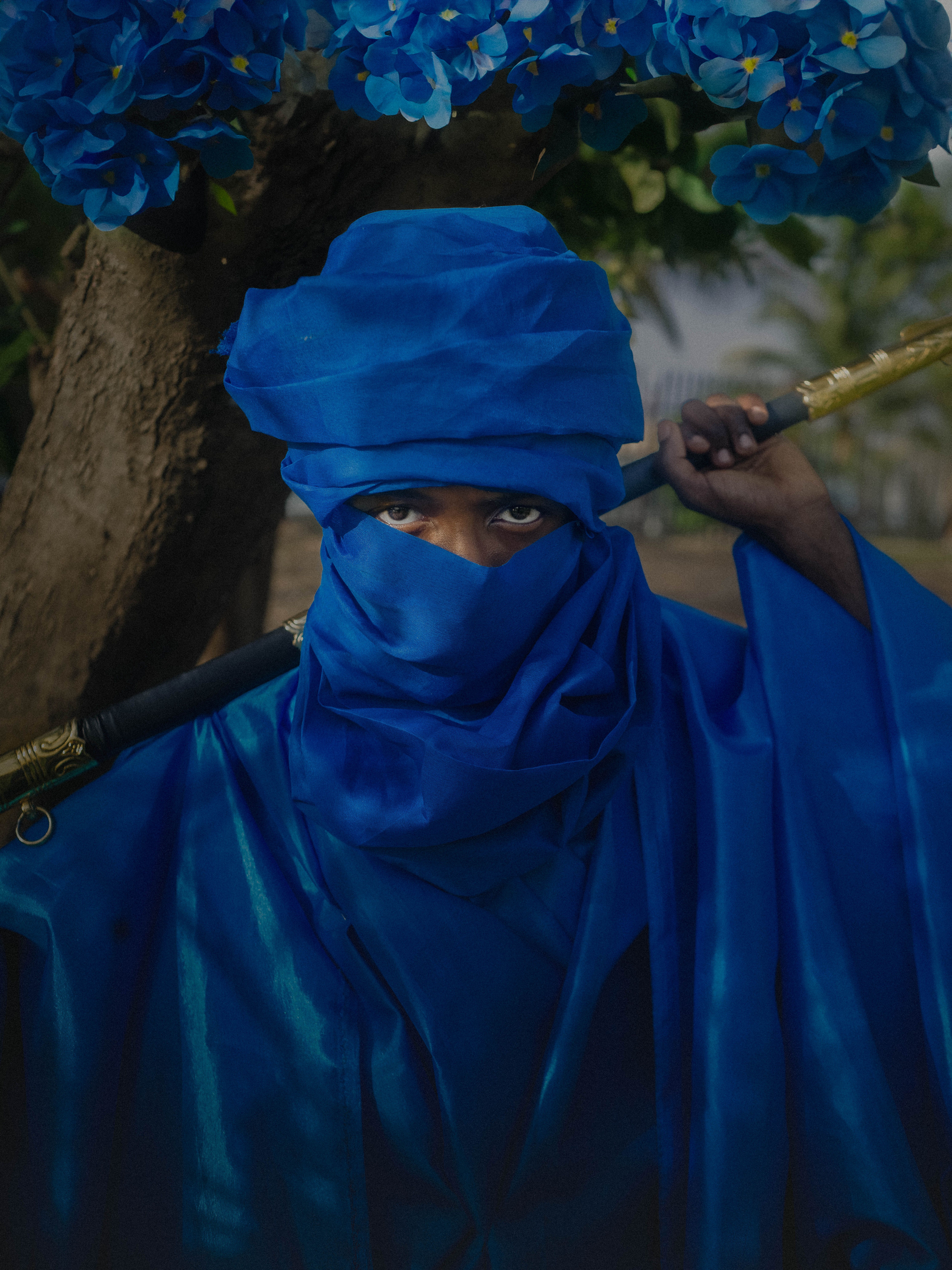 A person in traditional blue attire with a walking stick, outdoors in Abuja, Nigeria.