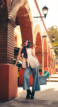A stylish woman poses confidently under a brick archway with outdoor ambient lighting and a vibrant fashion sense.