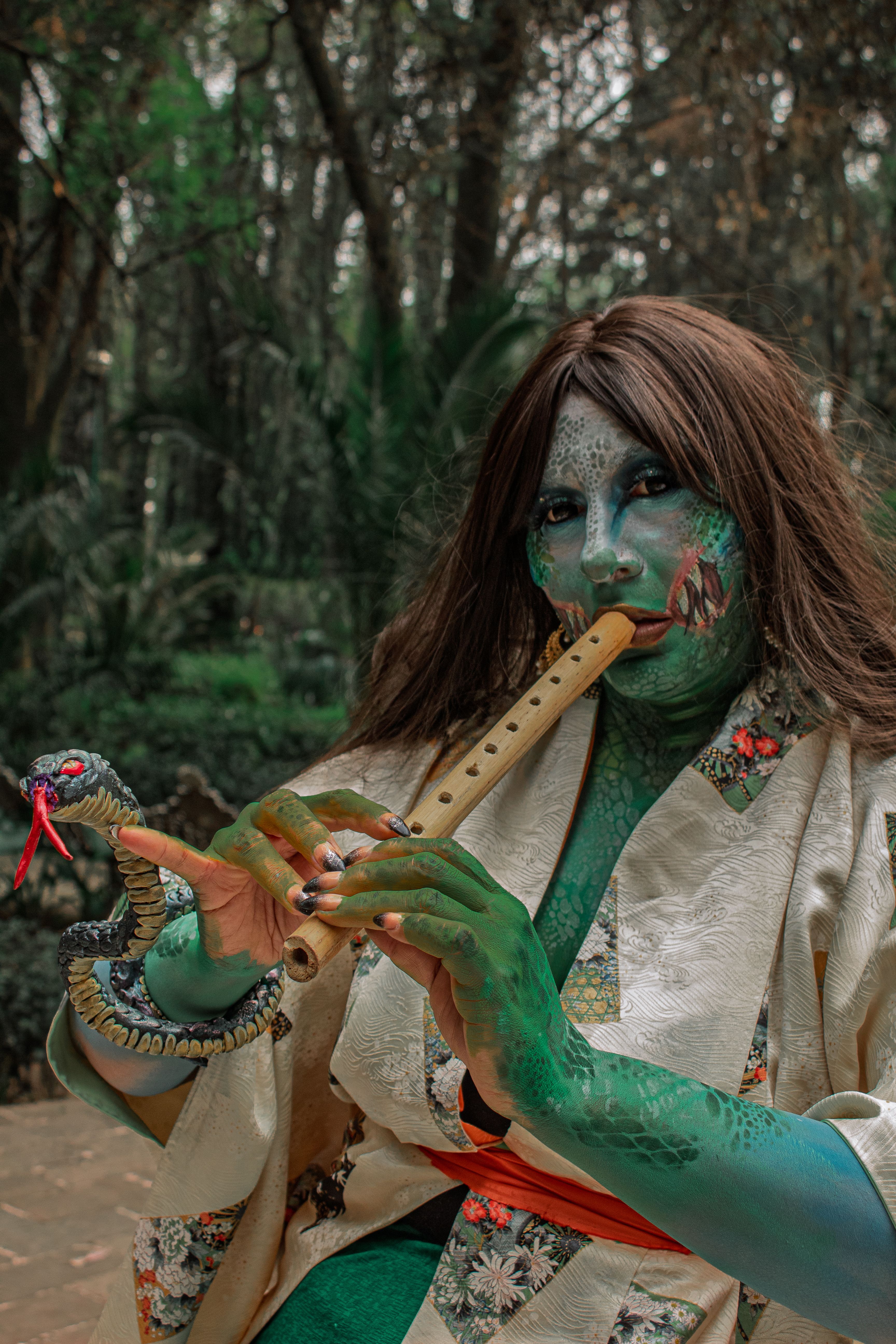 A person in fantasy costume playing a flute while holding a snake in a forest setting.