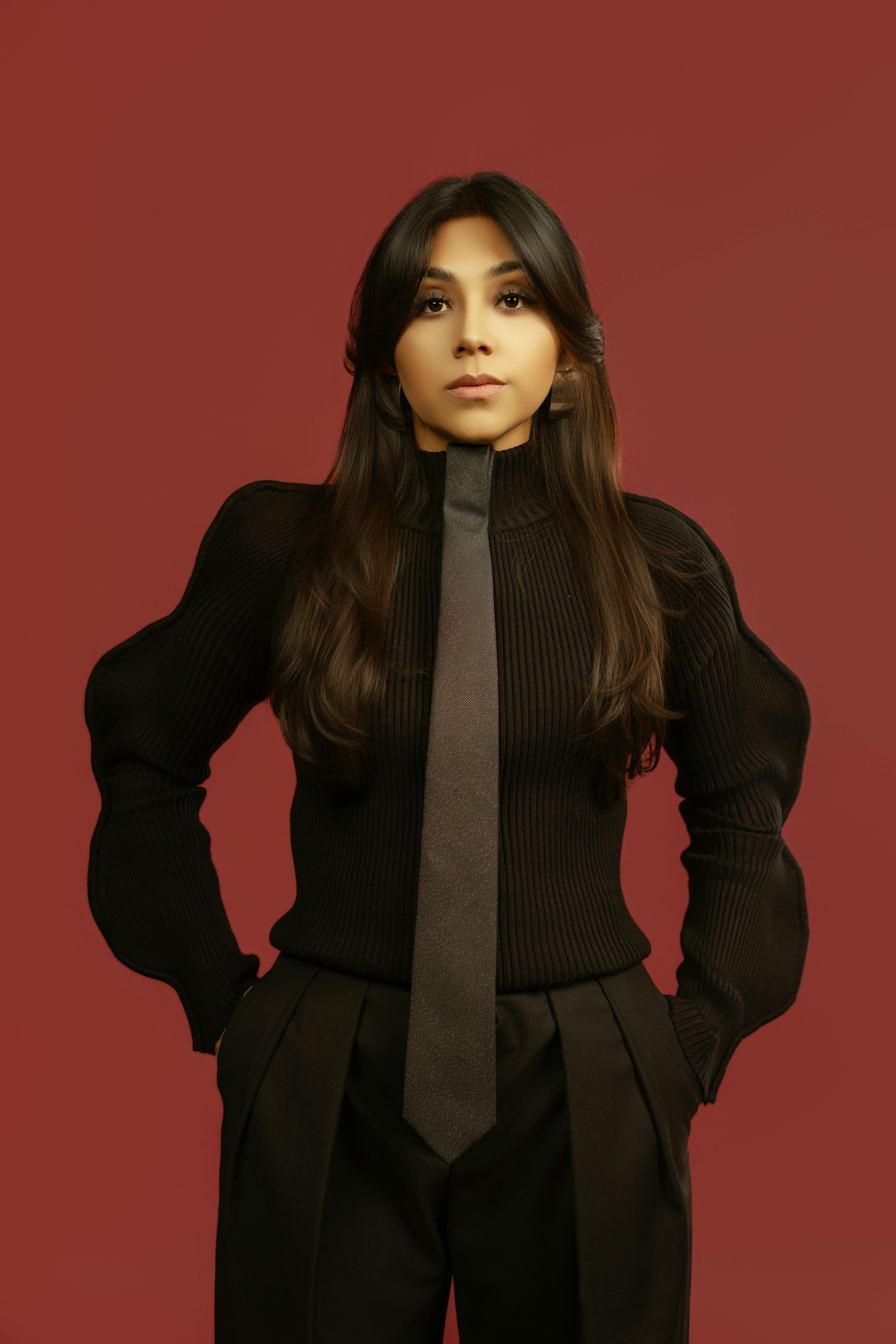 Free Portrait of a woman posing confidently wearing a black outfit and tie against a red background. Stock Photo