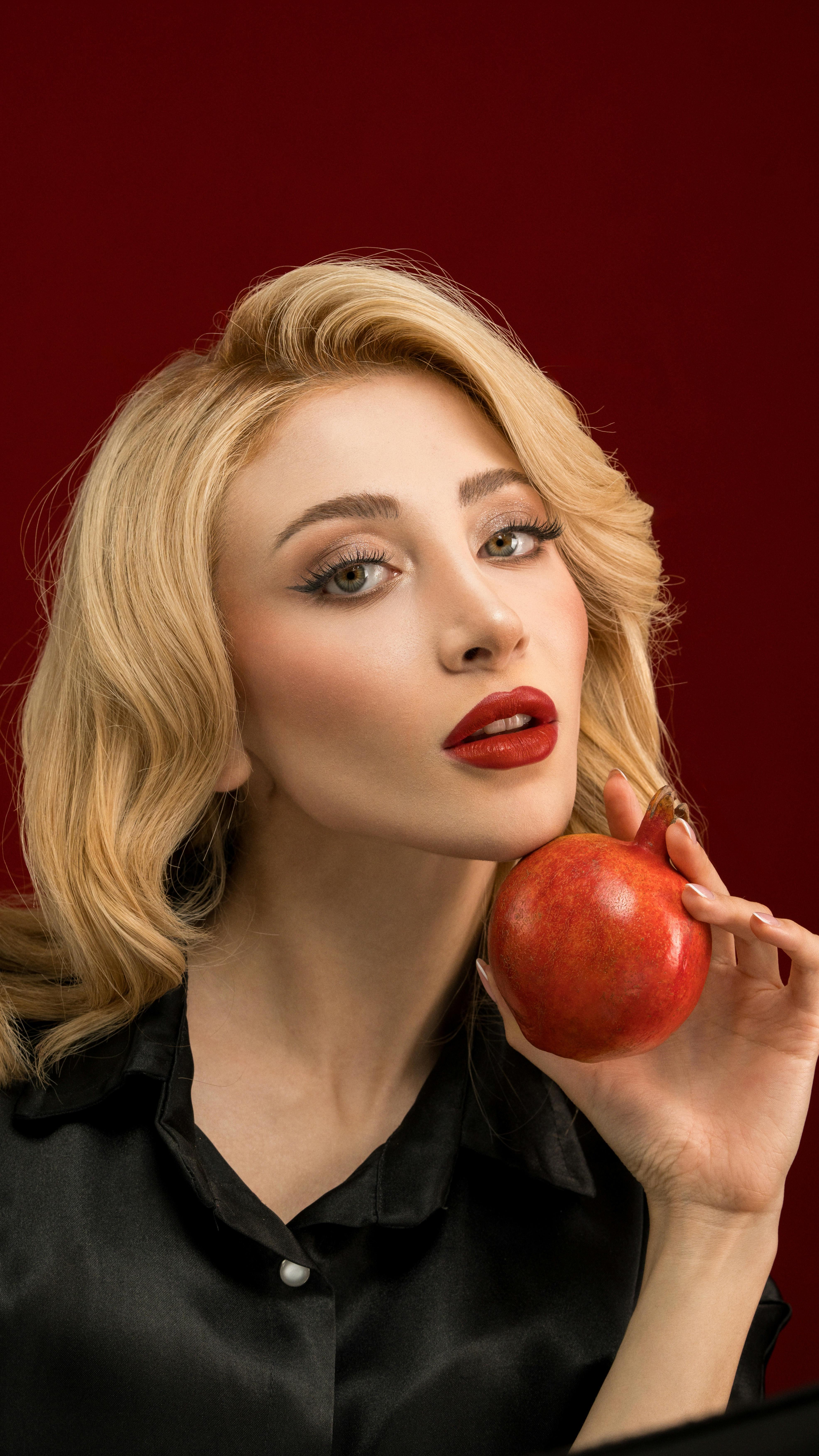 Glamorous portrait of a woman with red lips holding a red apple against a deep red background.