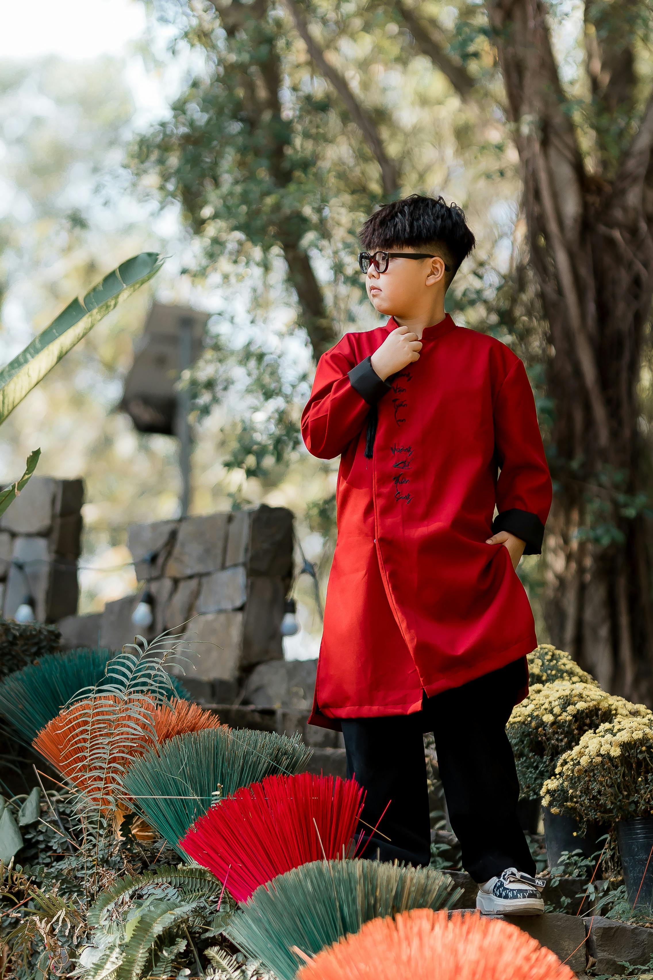 Boy in red traditional outfit standing outdoors amidst colorful fans and foliage.