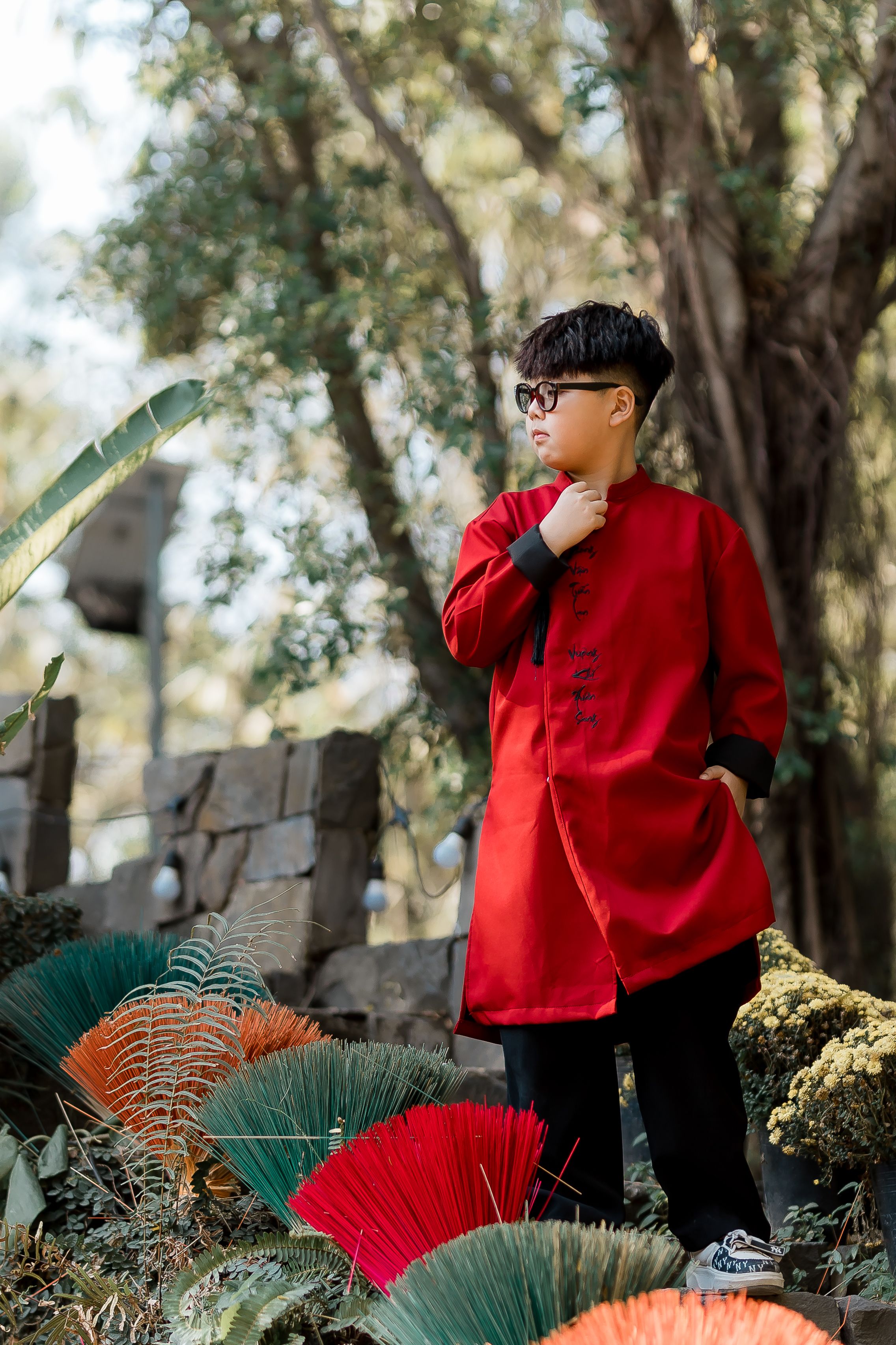 Young boy wearing traditional red attire stands outdoors surrounded by nature.