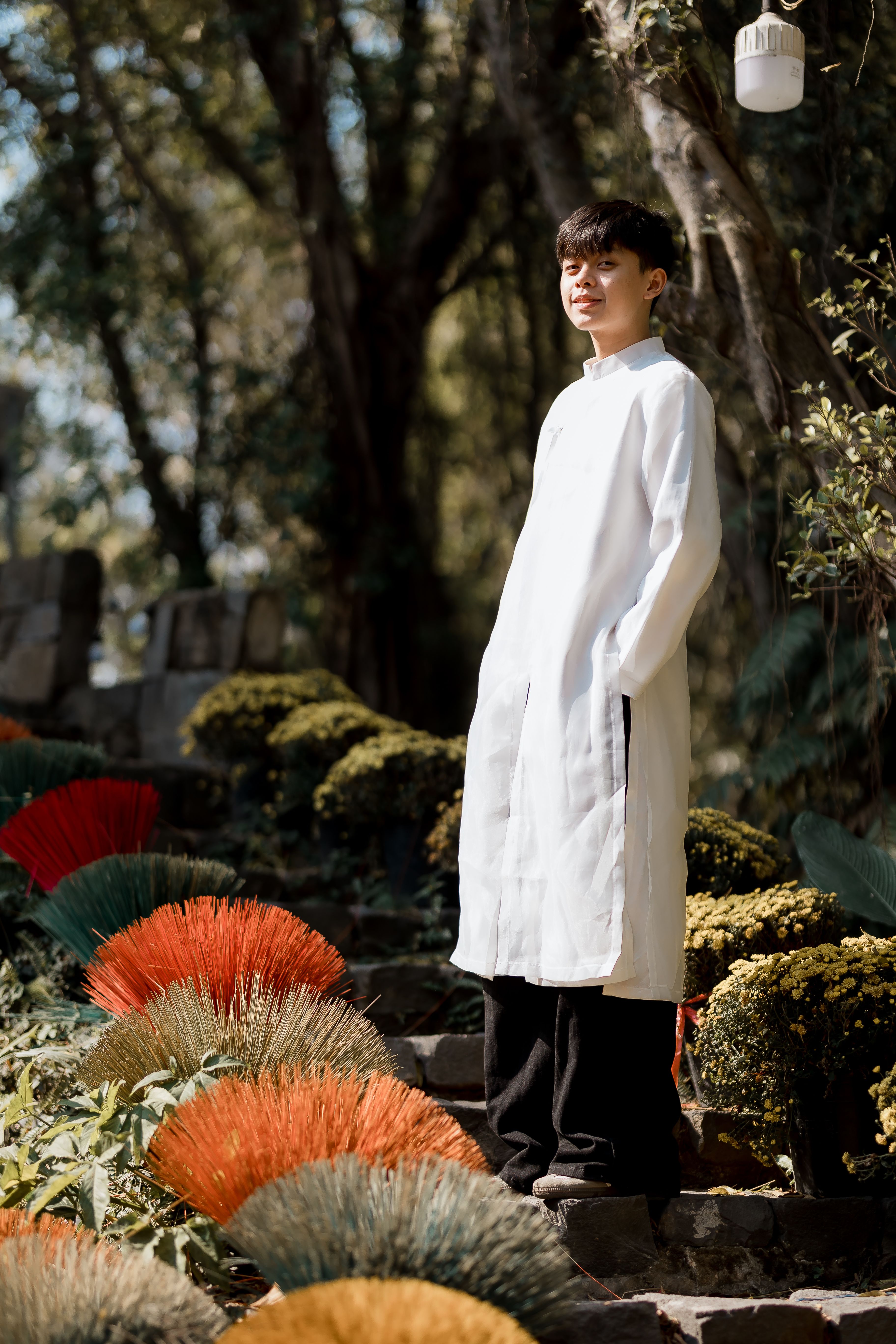 A man stands in a garden wearing traditional long white robe beside colorful flowers.