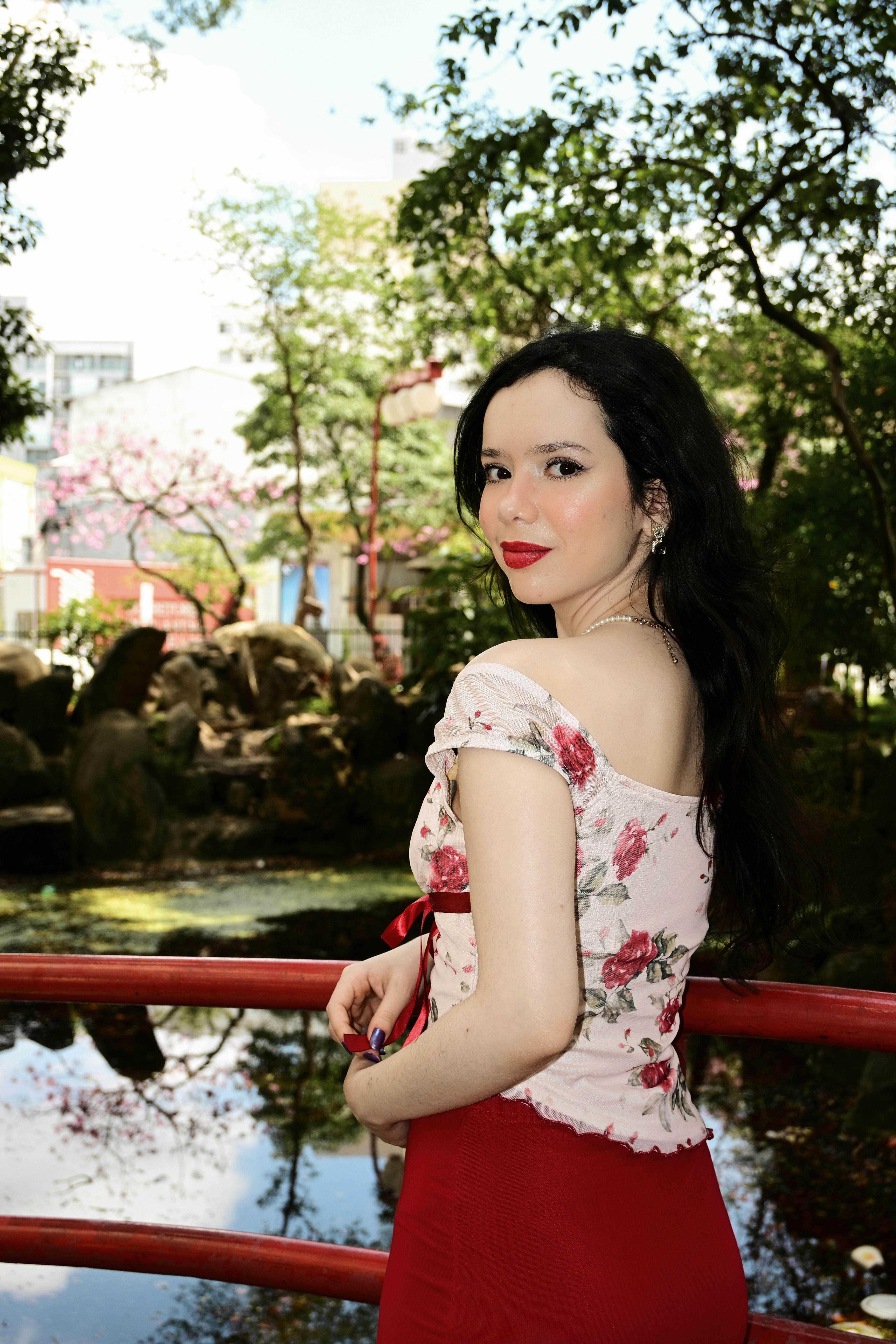 ฟรี หญิงสาวสวมเสื้อลายดอกไม้และกระโปรงสีแดงยืนอยู่ริมสระน้ำในสวนสาธารณะที่ล้อมรอบไปด้วยพืชพรรณเขียวชอุ่ม คลังภาพถ่าย