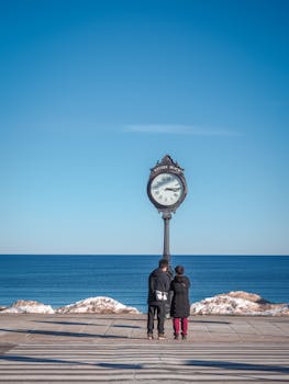 A couple stands by a clock on Revere Beach with the ocean and clear blue sky in the background.