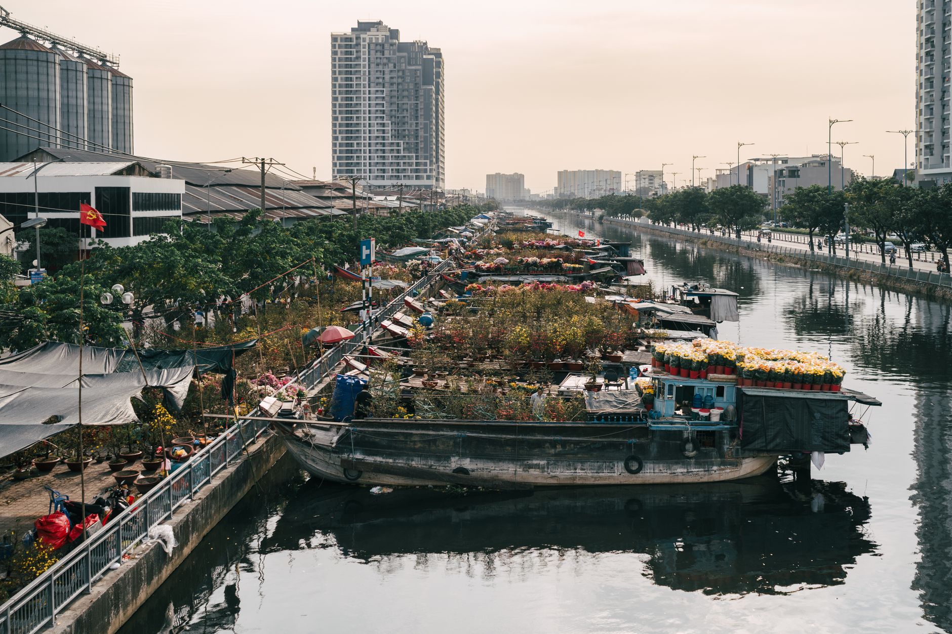 Flower boats line a calm river in a bustling city with towering buildings.