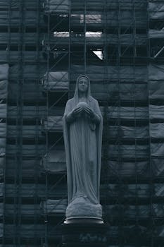Moody photo of Virgin Mary statue against a scaffolded building, mysterious ambiance.
