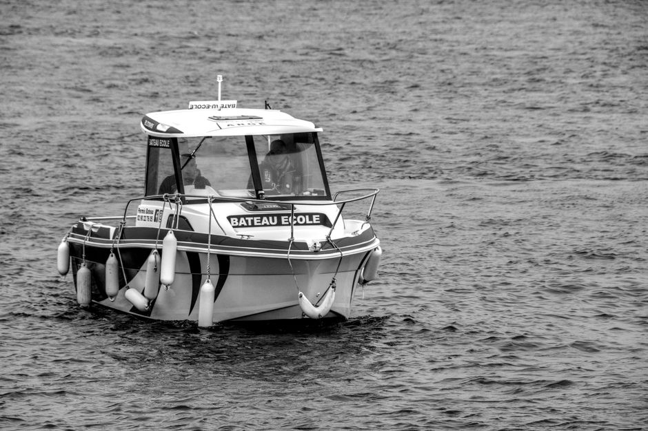 A black and white image showing a training boat labeled 'Bateau Ecole' on a calm sea.