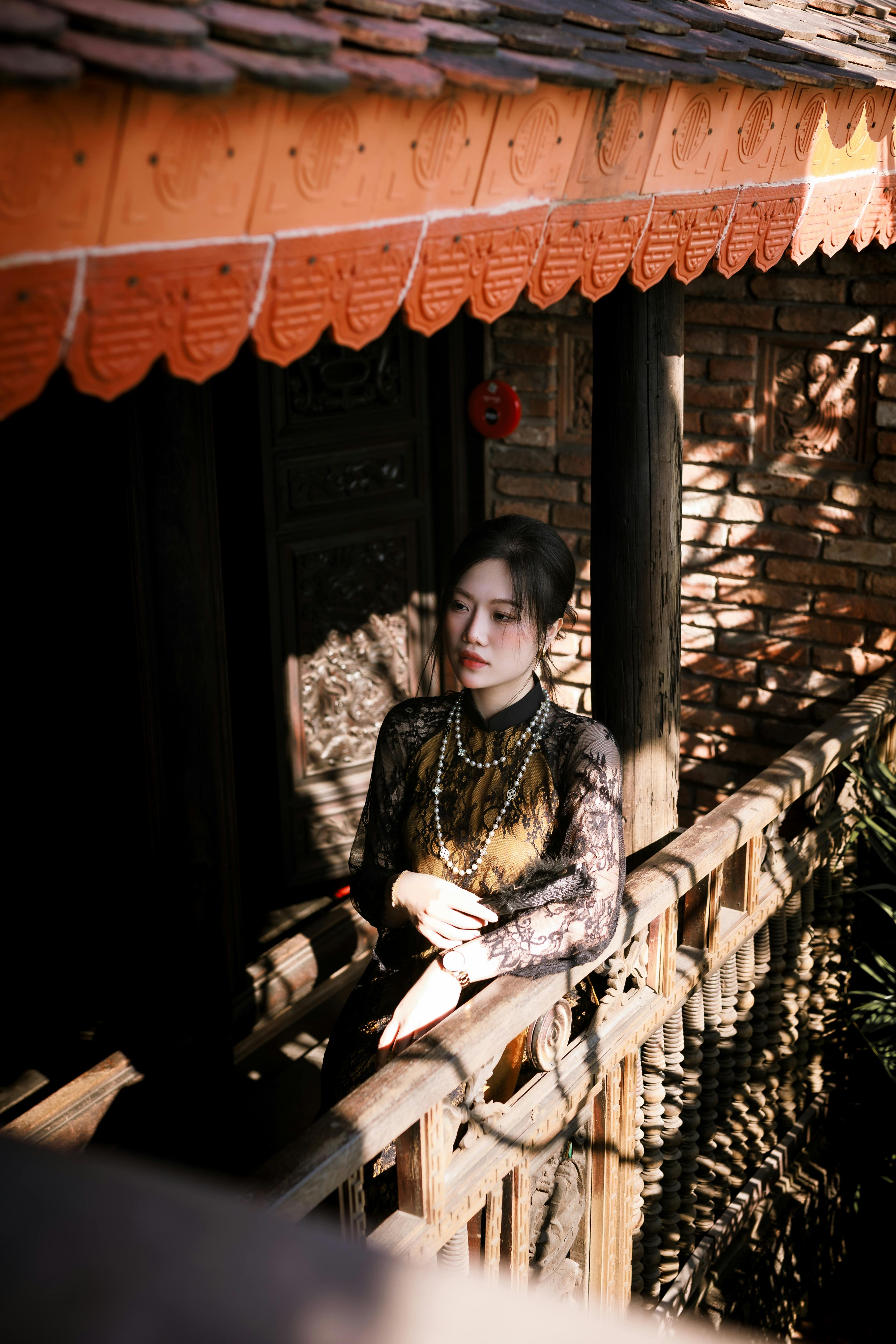 Gratis Una donna in abiti tradizionali è in piedi su un soleggiato balcone di legno, tipico dell'architettura asiatica. Foto a disposizione