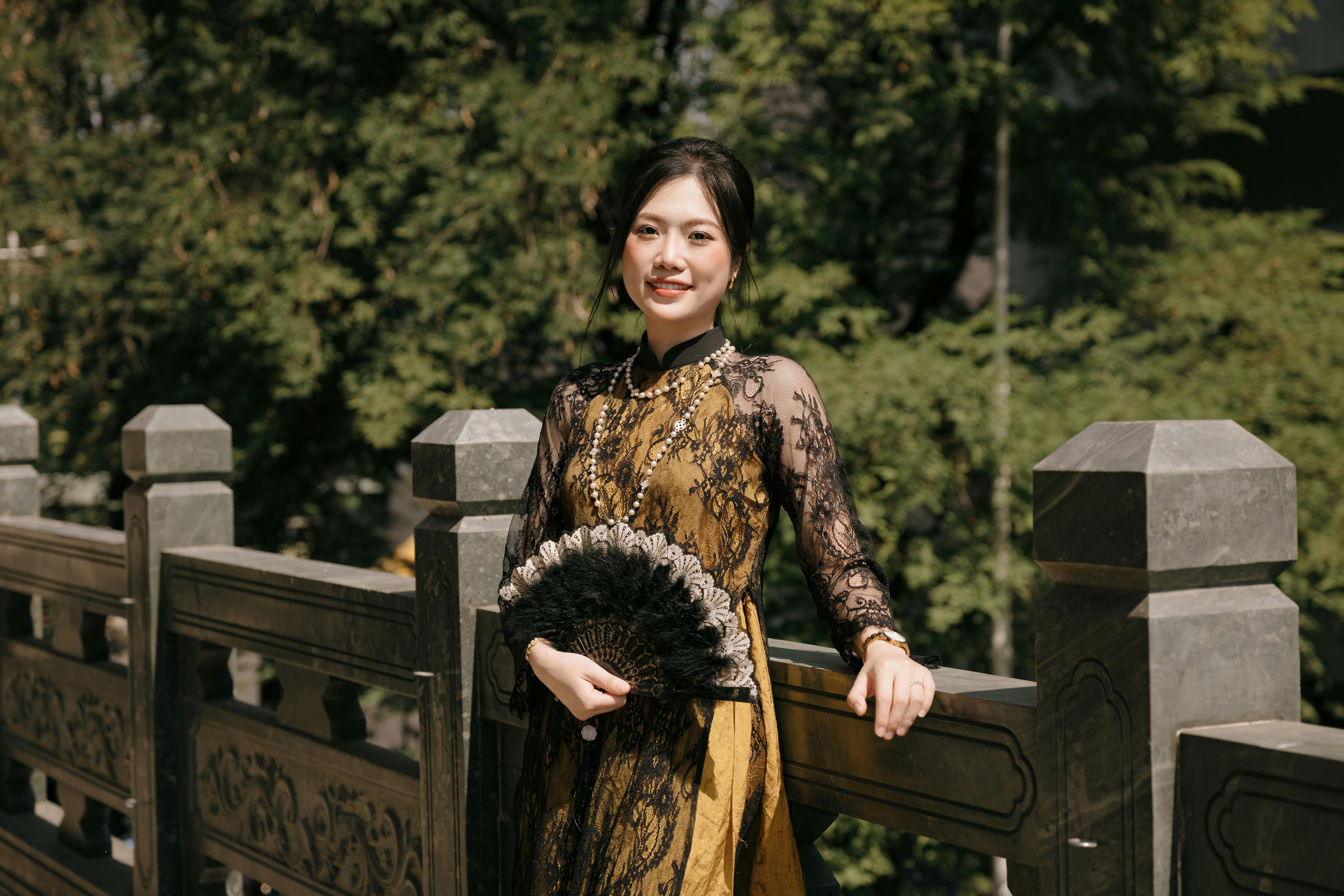 ฟรี หญิงสาวในชุดพื้นเมืองยืนอย่างสง่างามบนสะพานหิน ถือพัดประดับตกแต่งอยู่กลางแจ้ง คลังภาพถ่าย