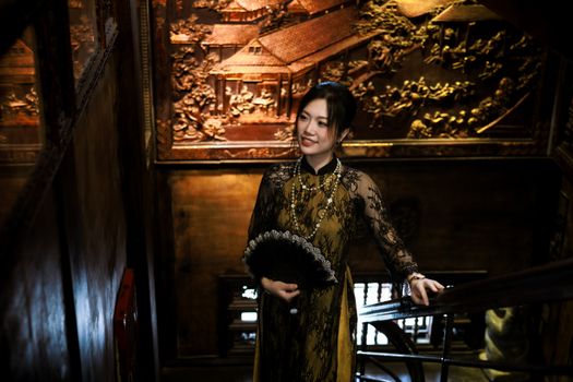 A woman in traditional Asian attire poses indoors with ornate wooden carvings in the background.