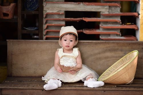 Free Adorable baby in a beige outfit holding a fan, seated with a basket hat nearby. Stock Photo