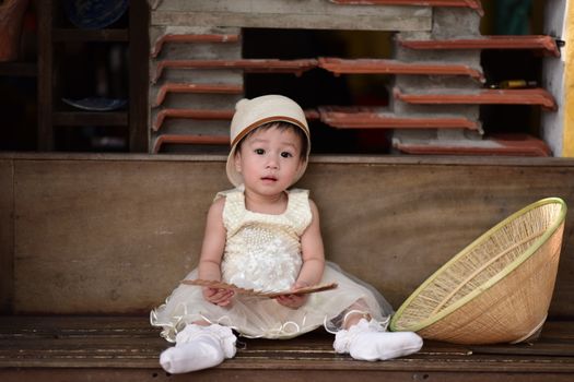 Cute baby in a dress sitting indoors with a bamboo hat, looking curious.