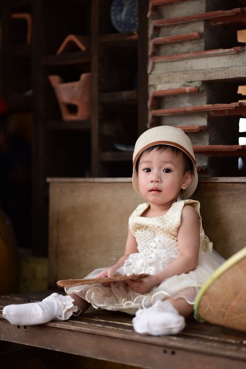 Free Adorable toddler in a vintage dress and hat, sitting indoors with soft lighting. Stock Photo