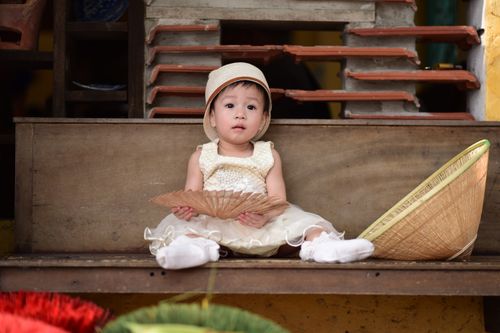Free Young child in vintage attire holding a hand fan, sitting in a rustic environment with traditional elements. Stock Photo
