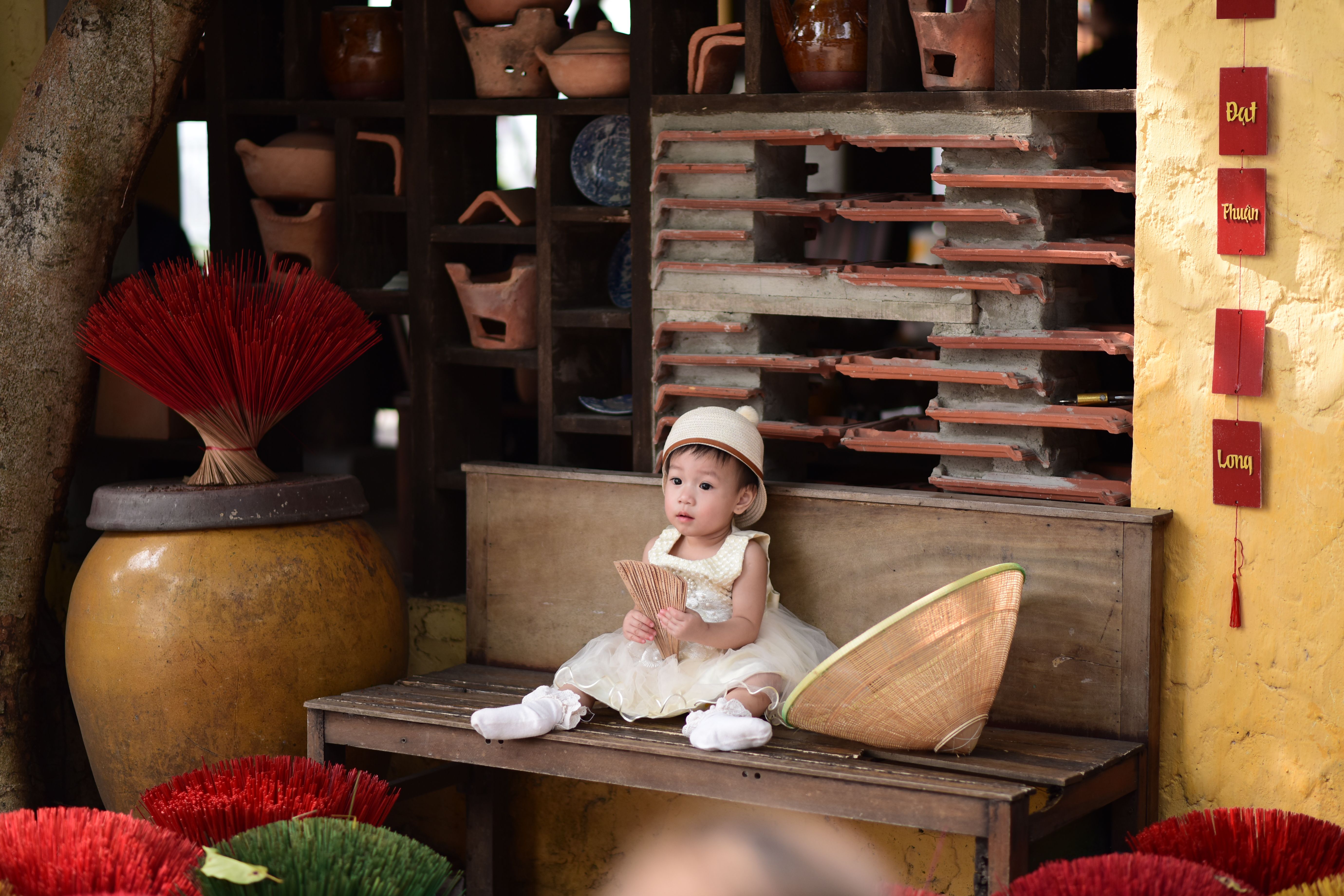 Adorable toddler wearing a dress and hat sits indoors with rustic decor, holding a fan.