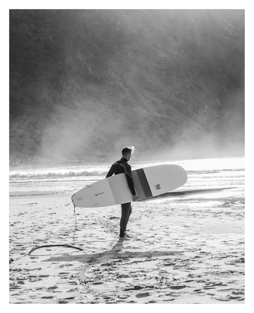 Grayscale Photo of Man Holding Surfboard Walking On Beach